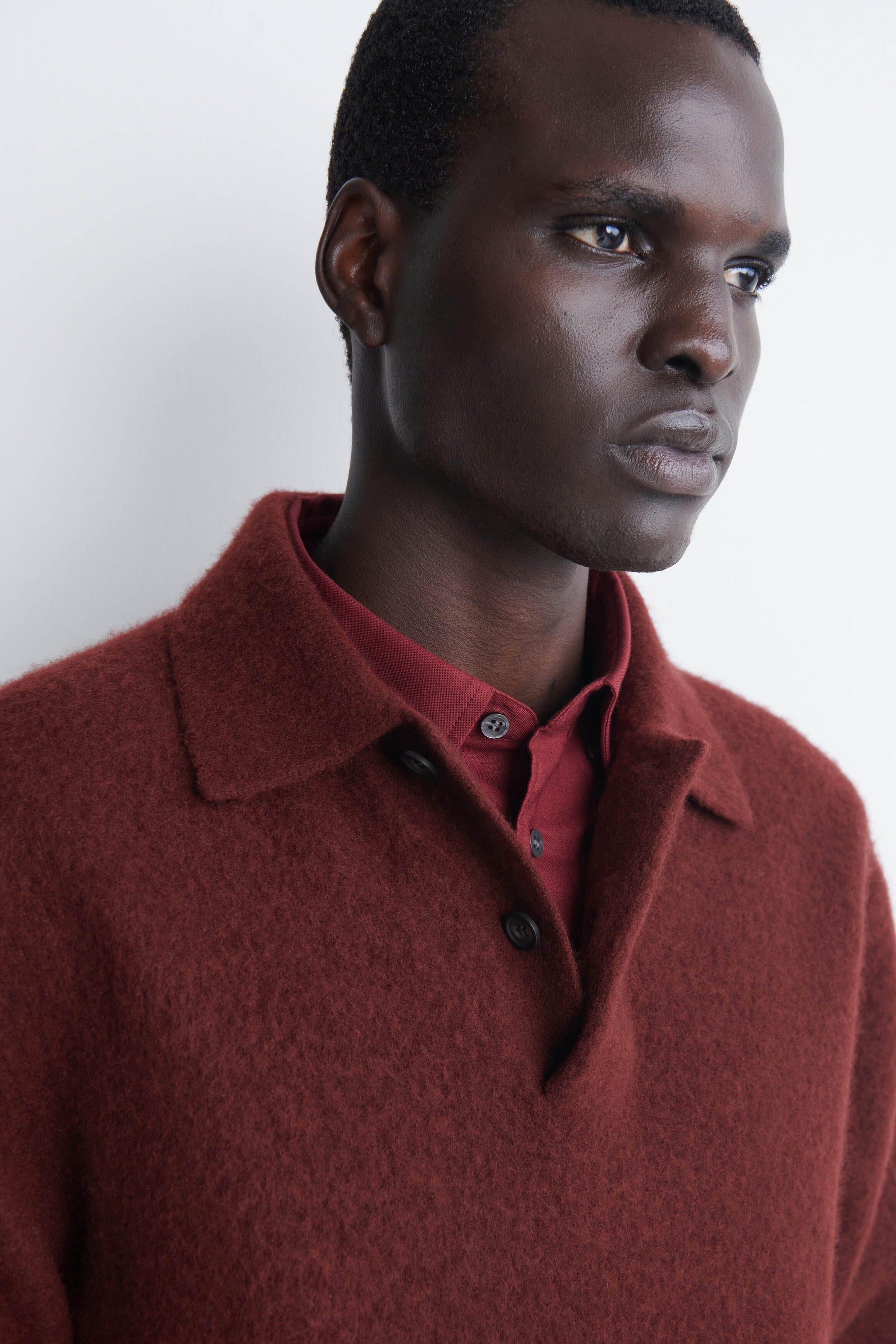 A rust-red brushed-wool polo shirt is shown from a front three-quarter angle, featuring a fuzzy, textured finish, a classic collar, and a partially open button placket. The polo shirt is layered over a smooth, deep red collared shirt. A rust-red brushed-wool polo shirt is shown from a front three-quarter angle, featuring a fuzzy, textured finish, a classic collar, and a partially open button placket. The polo shirt is layered over a smooth, deep red collared shirt.