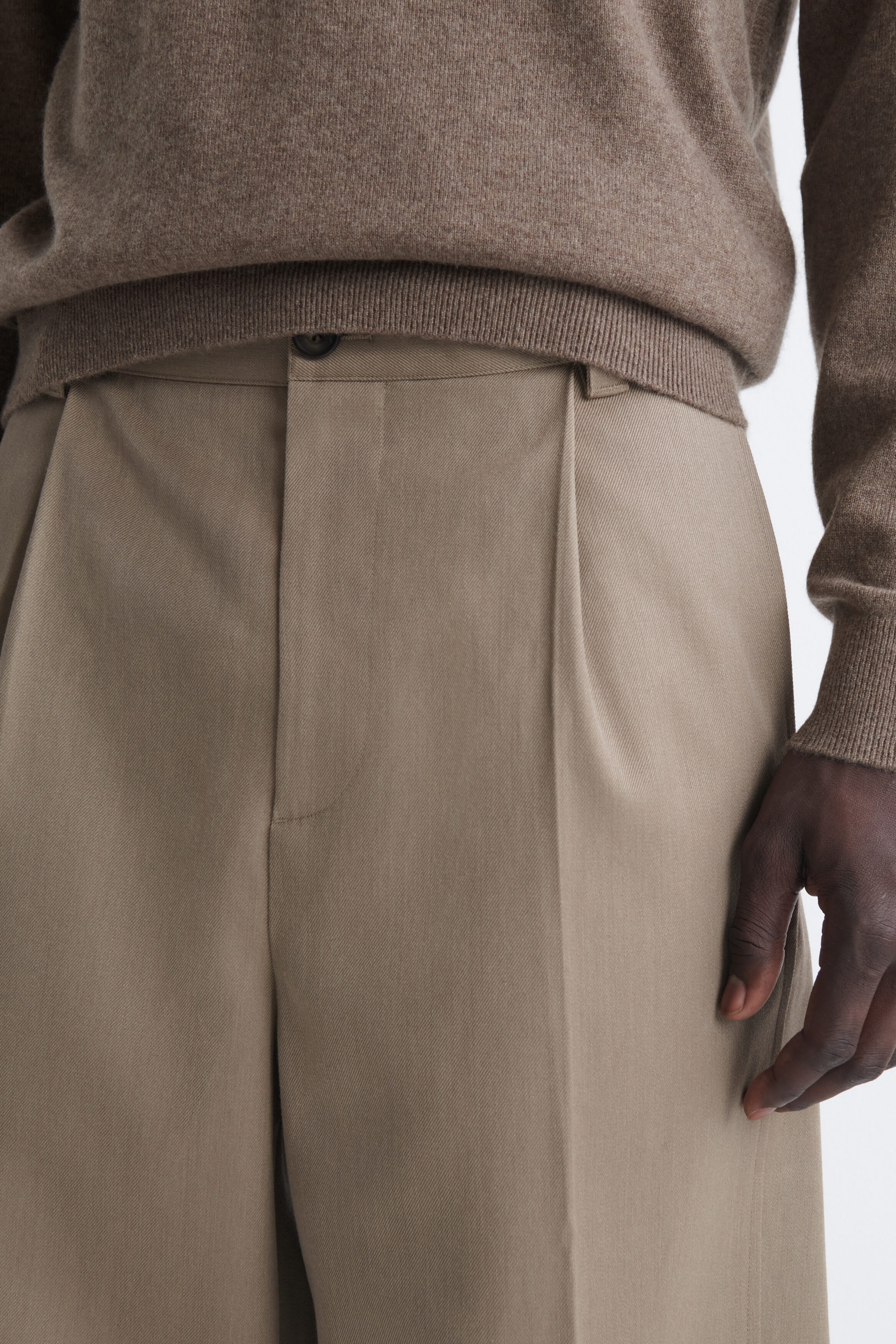 Estos pantalones beige con pinzas, vistos de frente en un primer plano, muestran una única pinza delantera, una cintura con cierre de botón y trabillas para cinturón, y una raya planchada a lo largo de la pernera. Un jersey de punto marrón con bajo acanalado descansa justo por encima de la cintura, dejando ver el detalle del botón superior. Estos pantalones beige con pinzas, vistos de frente en un primer plano, muestran una única pinza delantera, una cintura con cierre de botón y trabillas para cinturón, y una raya planchada a lo largo de la pernera. Un jersey de punto marrón con bajo acanalado descansa justo por encima de la cintura, dejando ver el detalle del botón superior.