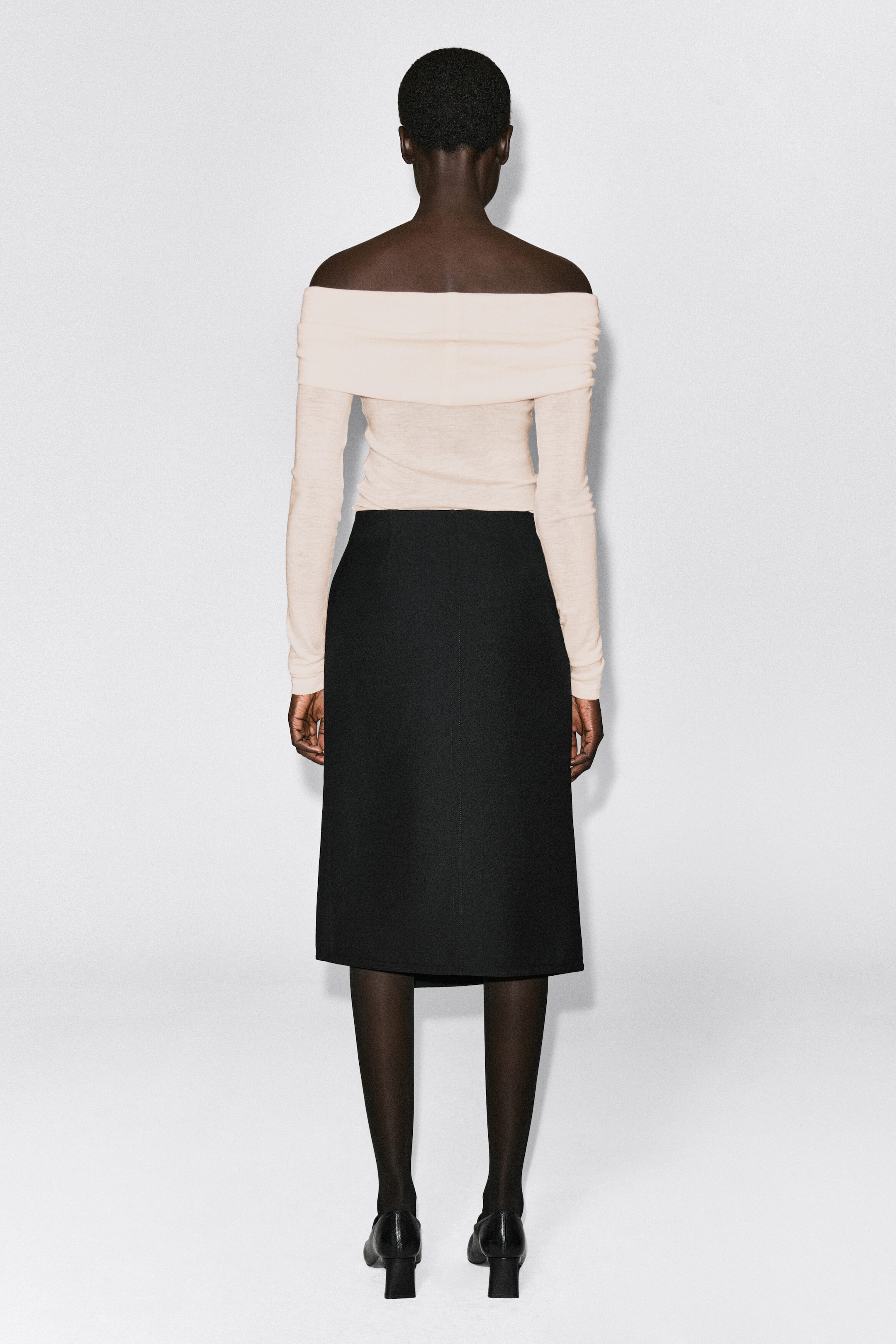 An off-the-shoulder merino wool top in a light cream hue is shown from the back, featuring a wide, folded collar band and long, slim-fitting sleeves. The top is tucked into a high-waisted black midi skirt, highlighting its close-fitting silhouette. An off-the-shoulder merino wool top in a light cream hue is shown from the back, featuring a wide, folded collar band and long, slim-fitting sleeves. The top is tucked into a high-waisted black midi skirt, highlighting its close-fitting silhouette.