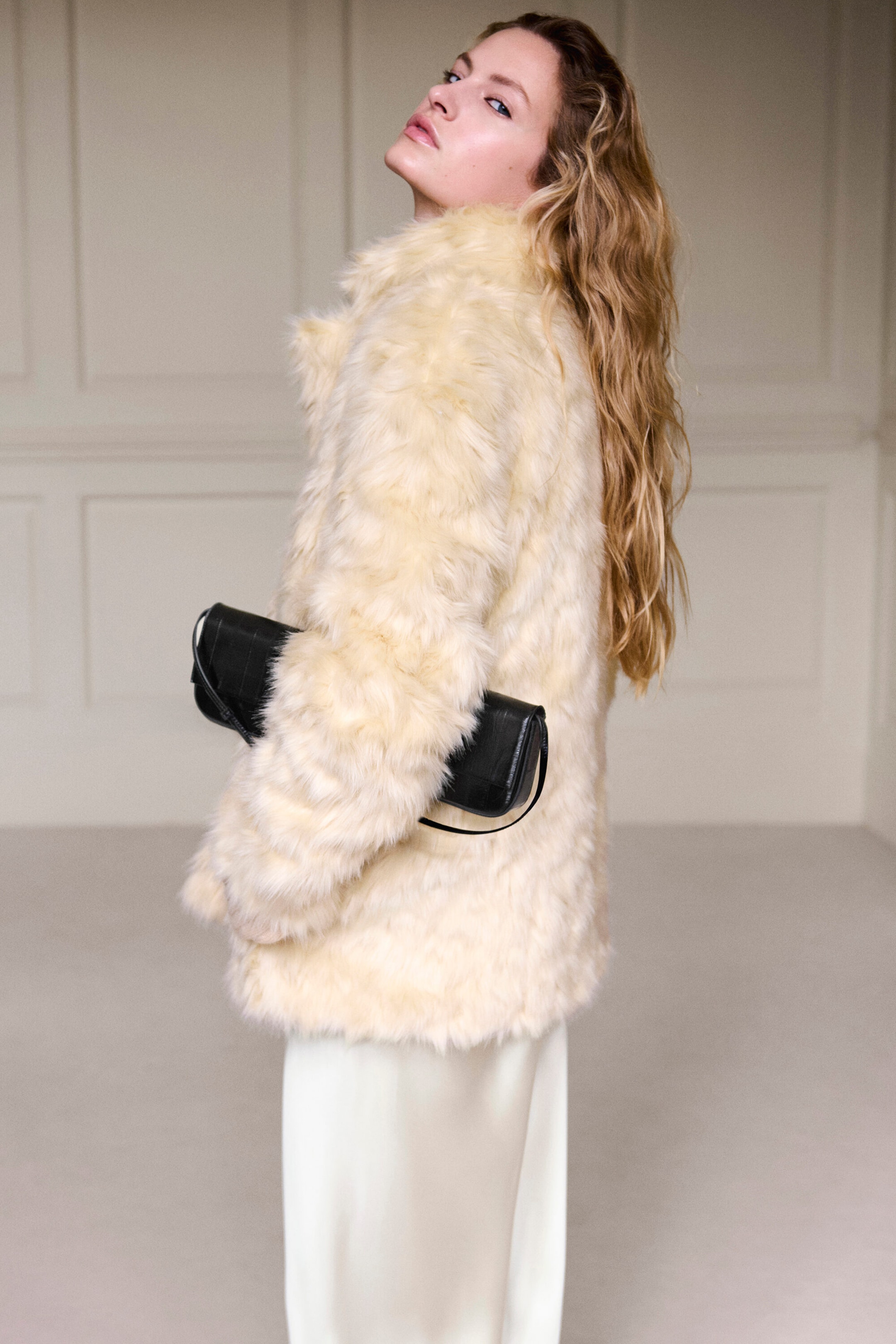 A cream-coloured fur-effect jacket is being shown from a three-quarter rear angle, highlighting its plush, shaggy faux-fur texture and relaxed fit. The hip-length jacket features long sleeves and is being styled with a black clutch held under the arm.