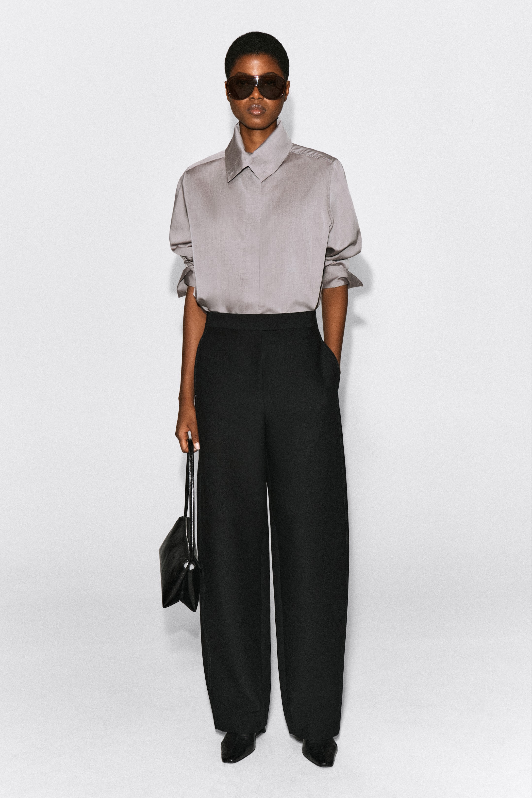 Agrandir l'image: Pantalon barrel noir à surpiqûres, taille haute et poches latérales, porté par le modèle avec une chemise grise boutonnée rentrée dans le pantalon. Le modèle est debout, une main posée dans la poche du pantalon, tout en tenant un petit sac à main en cuir noir.