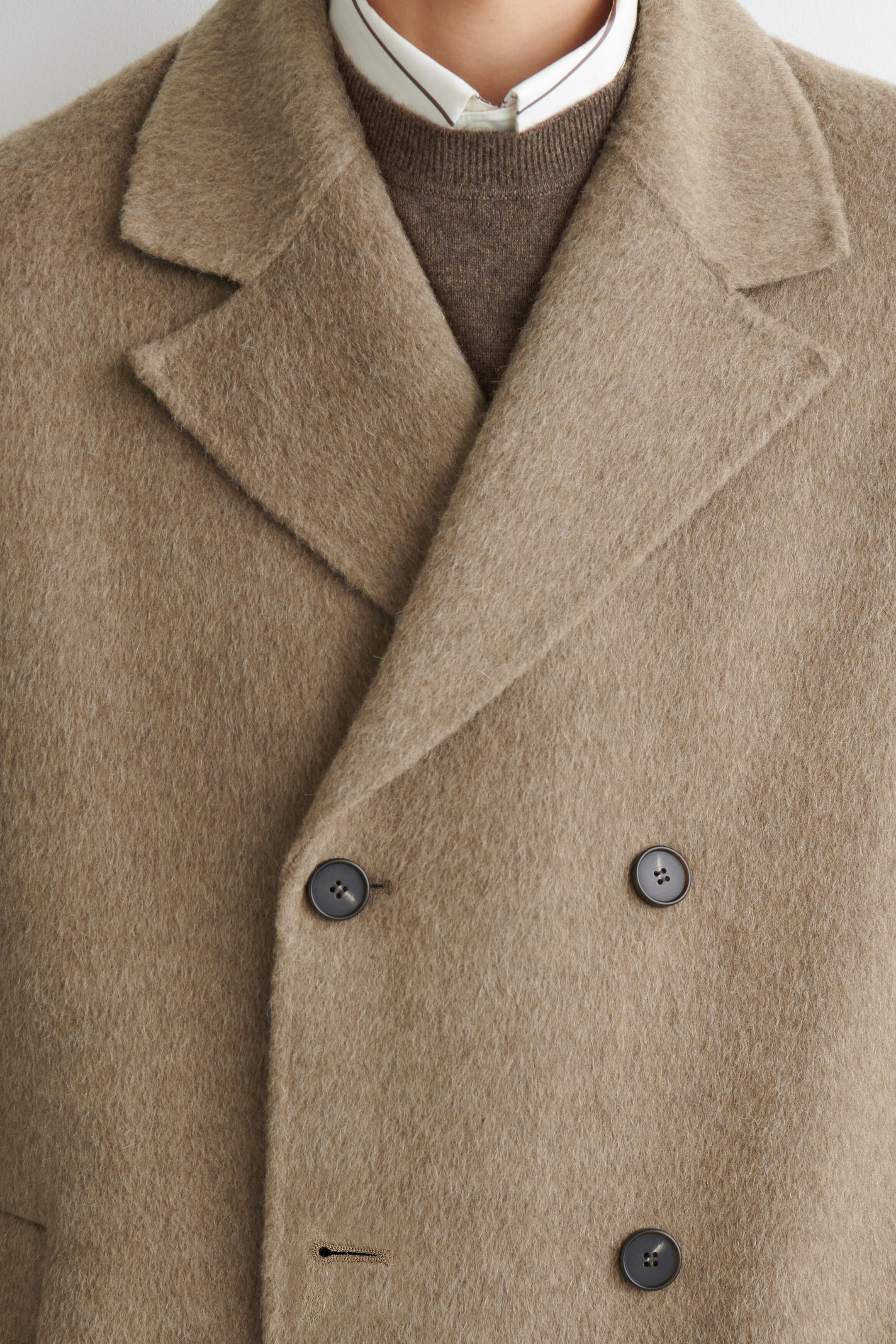 A close-up front view of a beige double-faced wool-blend long coat featuring wide notch lapels and a double-breasted front with dark buttons. The coat is layered over a brown crewneck sweater and a white collared shirt, highlighting its soft, brushed texture.