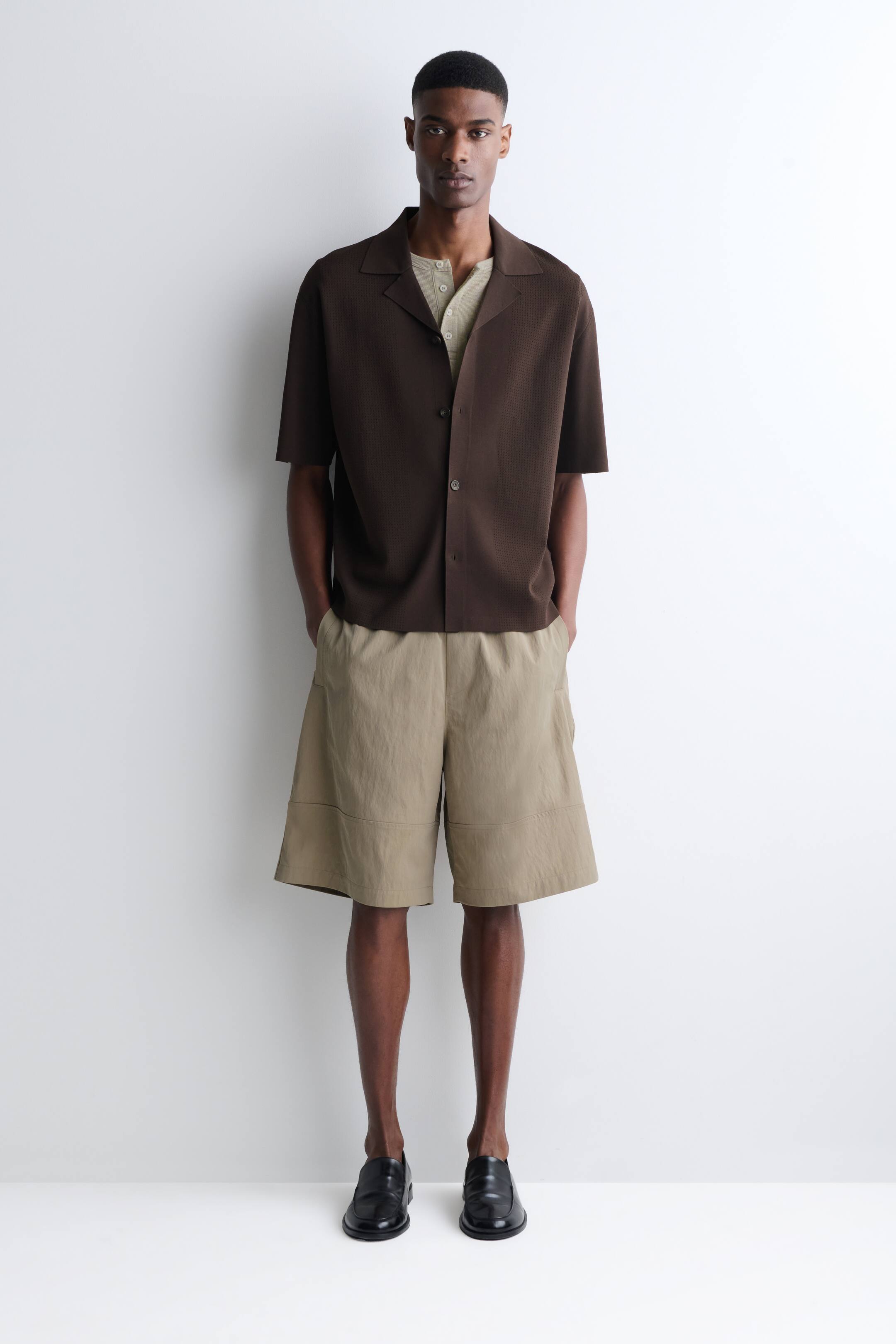Visualizza immagine più grande: Una camicia a maniche corte marrone scuro con una texture particolare e colletto a rever è indossata frontalmente, stratificata sopra una maglia Henley a maniche corte beige chiaro. Pantaloncini kaki a gamba larga con dettagli a pannello e mocassini neri slip-on completano l'outfit.