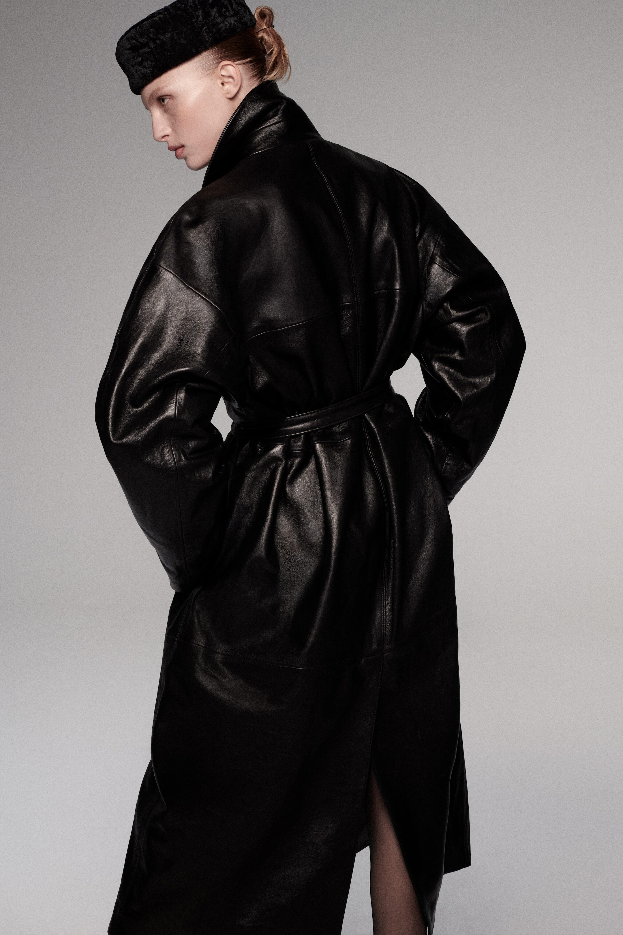 An oversized belted leather long coat is shown from a three-quarter back angle, highlighting its cinched waist with a matching leather belt and voluminous, dropped-shoulder sleeves. The coat features a high stand-up collar, paneling details across the back, and a center vent at the hem. An oversized belted leather long coat is shown from a three-quarter back angle, highlighting its cinched waist with a matching leather belt and voluminous, dropped-shoulder sleeves. The coat features a high stand-up collar, paneling details across the back, and a center vent at the hem.