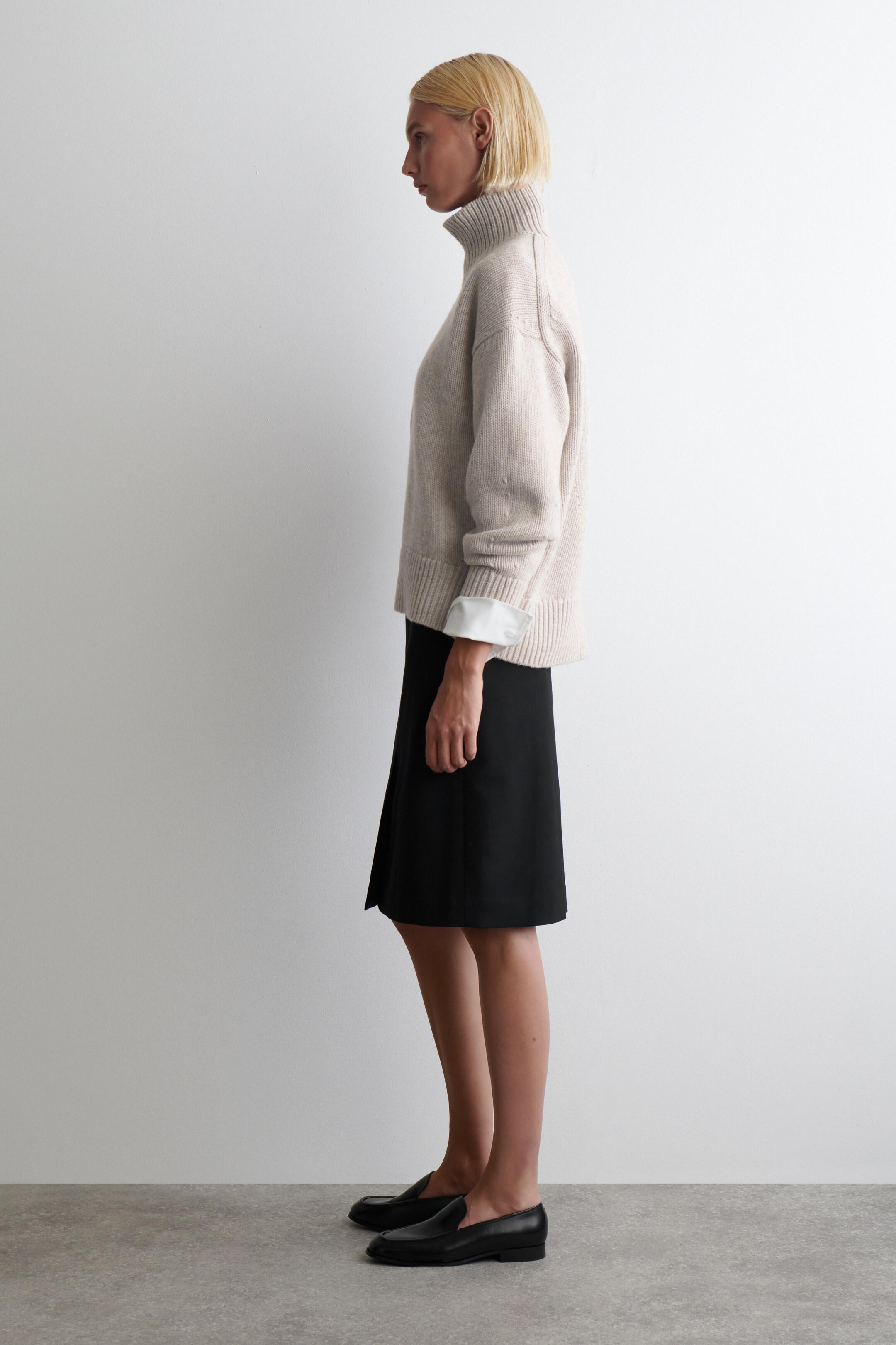 Grösseres Bild ansehen: Ein hellbeiger, locker sitzender Rollkragenpullover wird im Seitenprofil präsentiert, wobei der gerippte Kragen und der tiefe gerippte Saum hervorgehoben werden. Die Ärmel des Pullovers sind hochgekrempelt, wodurch die weißen Manschetten eines darunter getragenen Hemdes mit Kragen sichtbar werden. Ein schwarzer knielanger Rock und schwarze flache Loafer werden ebenfalls getragen.