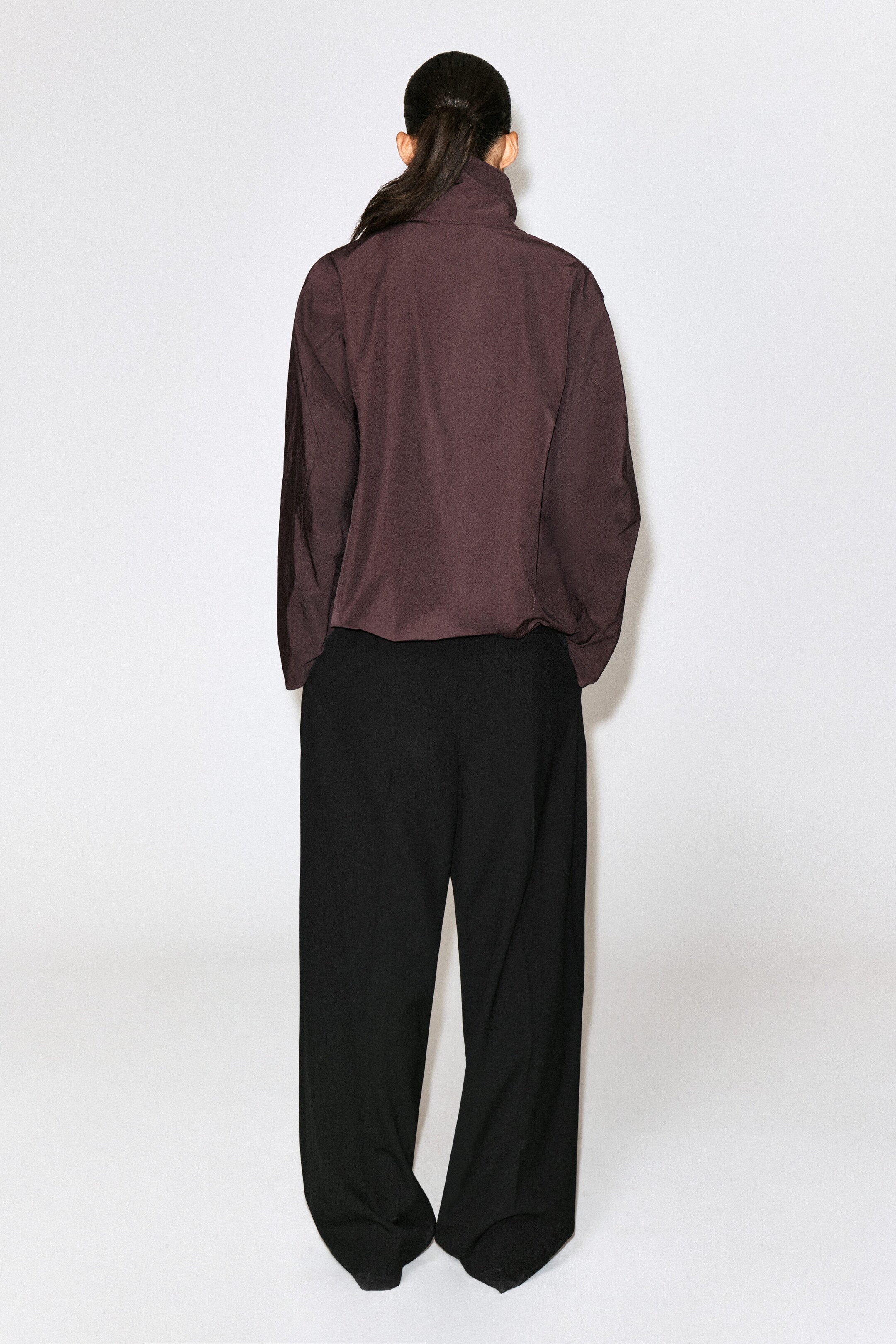 This dark plum-brown high-collar shirt is shown from the back, featuring long sleeves and a subtly gathered hem. It is worn with wide-leg black trousers to showcase its relaxed, structured drape. This dark plum-brown high-collar shirt is shown from the back, featuring long sleeves and a subtly gathered hem. It is worn with wide-leg black trousers to showcase its relaxed, structured drape.