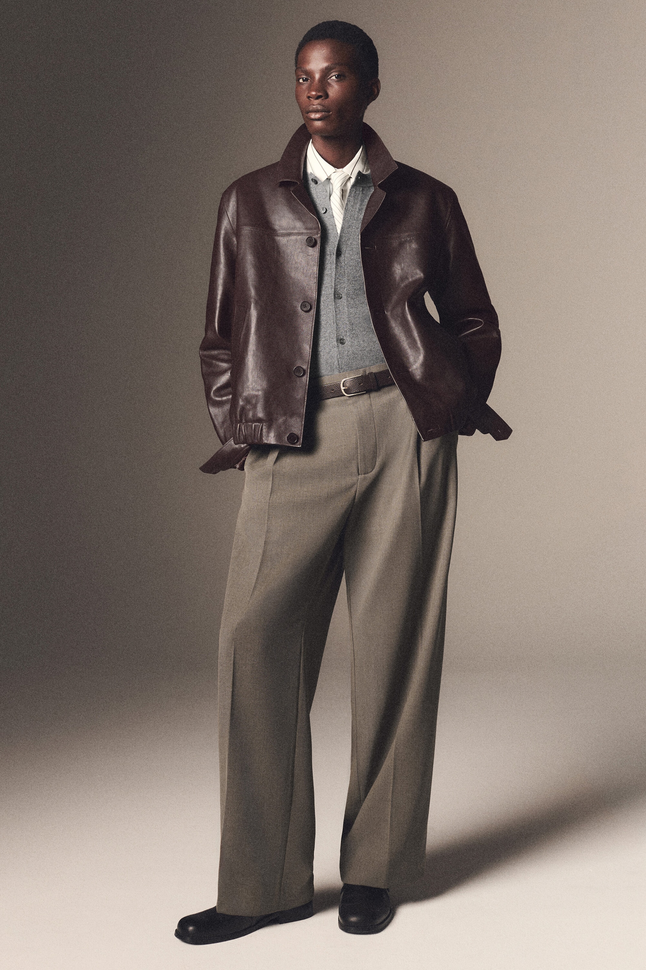 Un mannequin de face porte un blouson bomber en cuir marron foncé avec col pointu et ourlet élastiqué, superposé à un cardigan en maille gris et une chemise blanche à col. Il associe la veste à un pantalon large coupe décontractée en mélange laine taupe, mettant en valeur des plis devant et des plis marqués au centre des jambes. Un mannequin de face porte un blouson bomber en cuir marron foncé avec col pointu et ourlet élastiqué, superposé à un cardigan en maille gris et une chemise blanche à col. Il associe la veste à un pantalon large coupe décontractée en mélange laine taupe, mettant en valeur des plis devant et des plis marqués au centre des jambes.