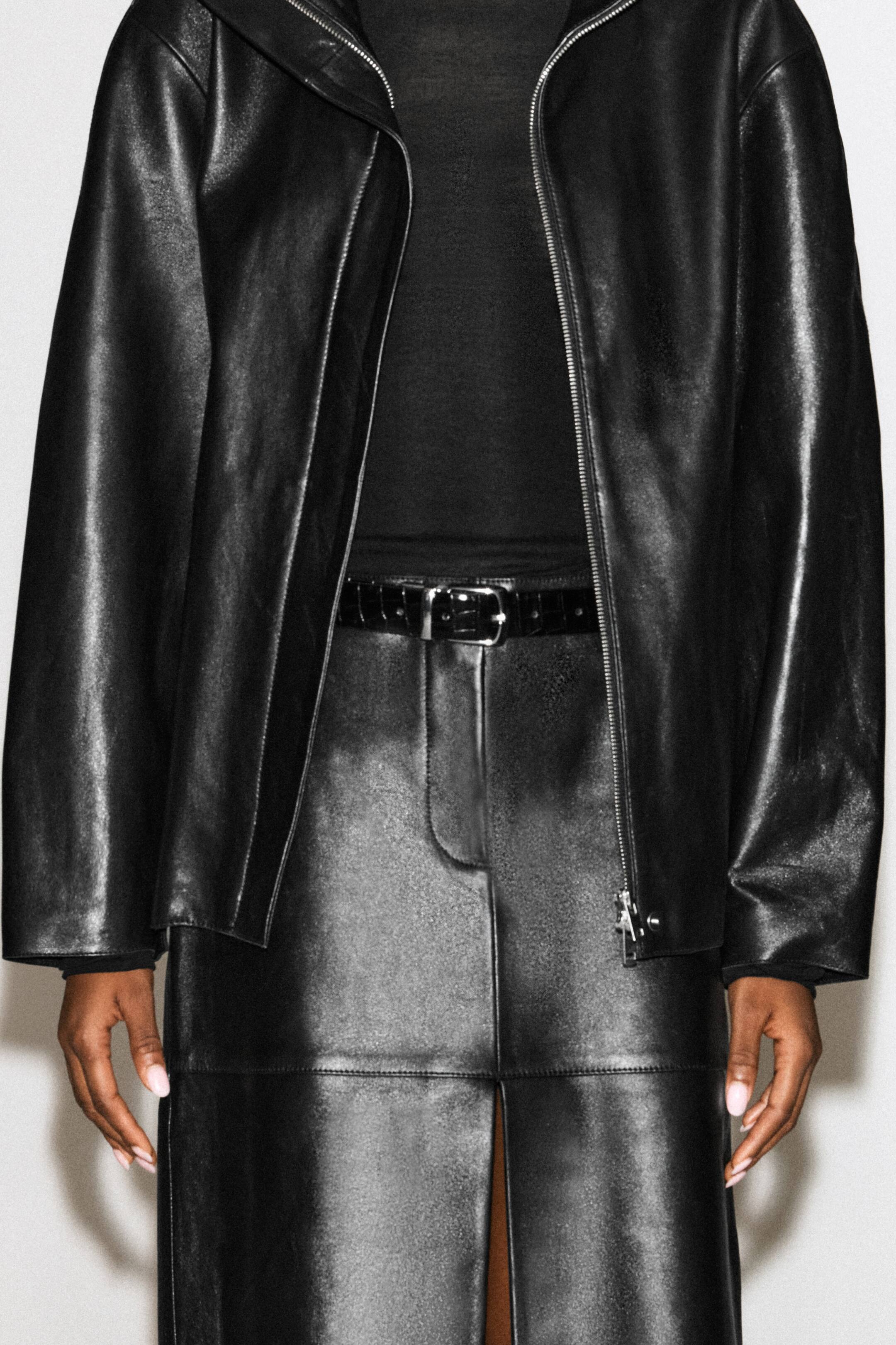 Grotere afbeelding bekijken: Een zwarte riem van leer met crocoprint en een zilverkleurige gesp wordt gedragen rond de taille van een zwarte leren rok. De riem is gestyled bij het model, dat ook een zwarte top en een open zwarte leren jas draagt.