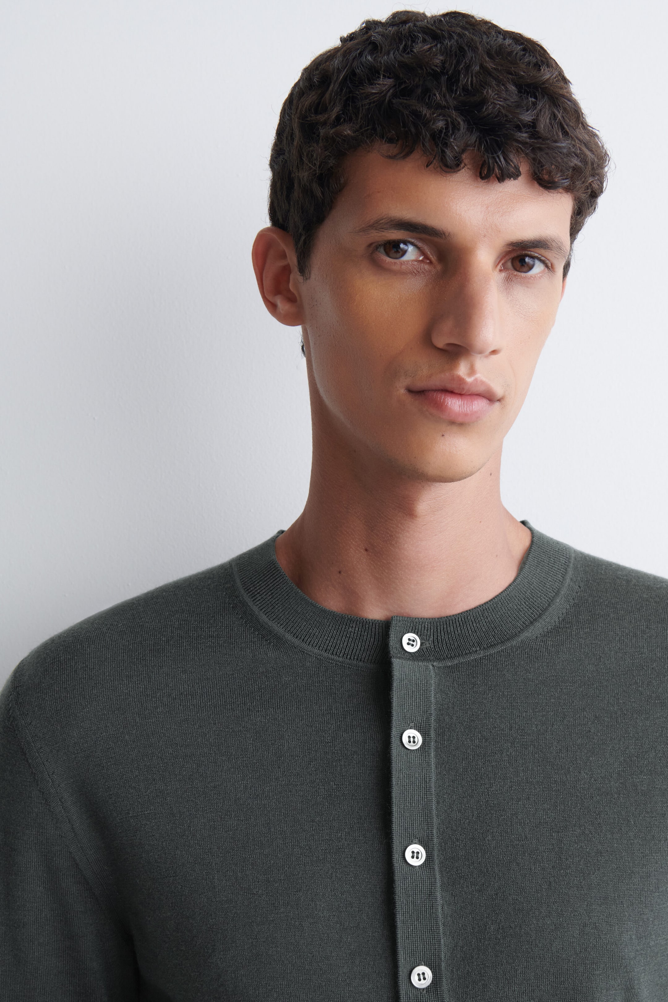 The model is wearing a dark green slim-fit merino wool Henley top. The fine-knit top features a ribbed crew neckline and a vertical front placket with four contrasting light-coloured buttons.