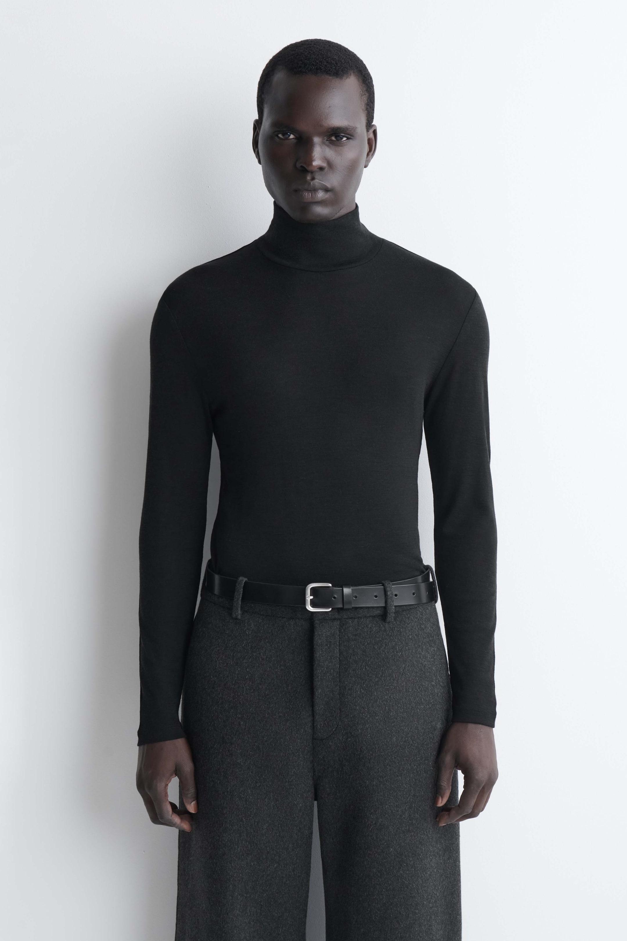 Größeres Bild anzeigen: Ein schmal geschnittenes Rollkragen-Top aus Wolljersey in Schwarz wird von vorne gezeigt und betont seinen hohen, umgeschlagenen Rollkragen sowie die körpernahe Silhouette. Das langärmelige Top ist in eine dunkelgraue Hose mit schwarzem Ledergürtel gesteckt und demonstriert dadurch seine leichte, glatte Textur.