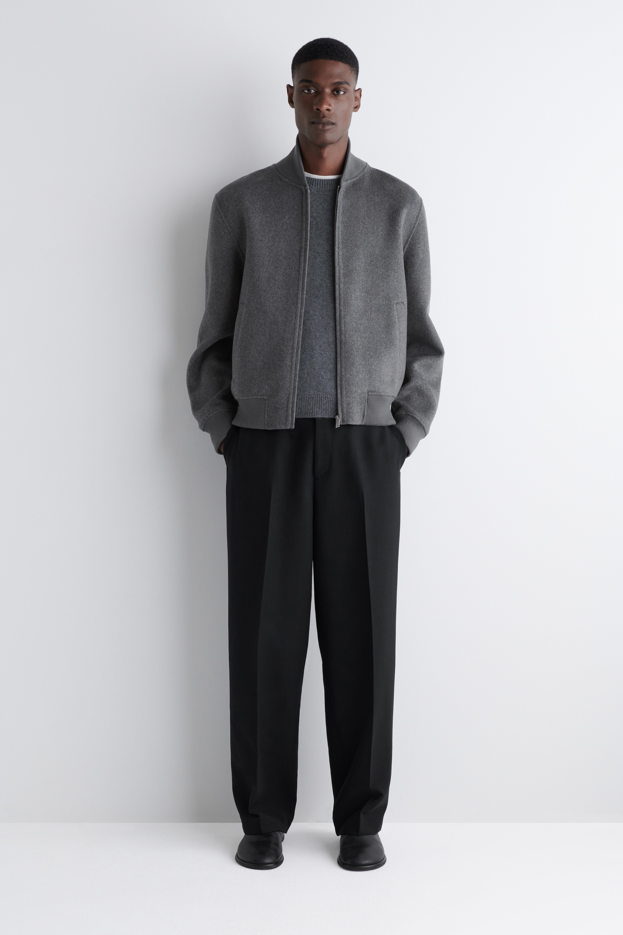 Ver imagen más grande: Una chaqueta bomber gris oscuro se presenta desde una vista frontal, mostrando su cuello alto acanalado, puños acanalados y dobladillo acanalado, junto con un cierre completo con cremallera y bolsillos de ojal visibles. Está superpuesta sobre un suéter de cuello redondo gris oscuro. La chaqueta y el suéter se combinan con pantalones de pierna ancha negros con una raya al frente y zapatos de vestir negros.