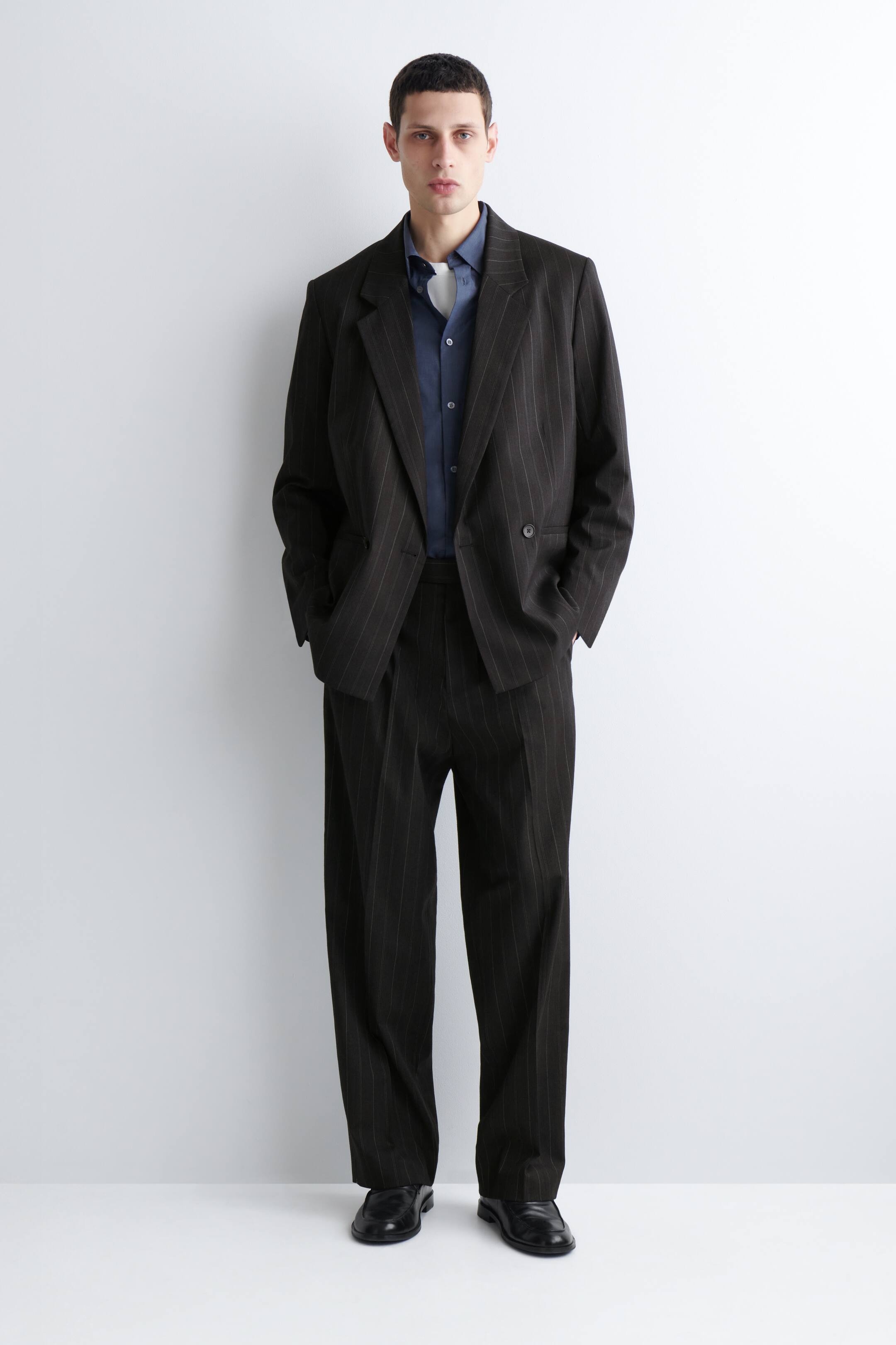 Преглед на по-голямо изображение: Представен е костюм с предно разположение в тъмно кафяво с райета, състоящ се от двуредно сако с назъбени ревери и подходящи панталони с широки крачоли. Под него е наслоена тъмносиня риза с яка, а черни официални обувки завършват ансамбъла.