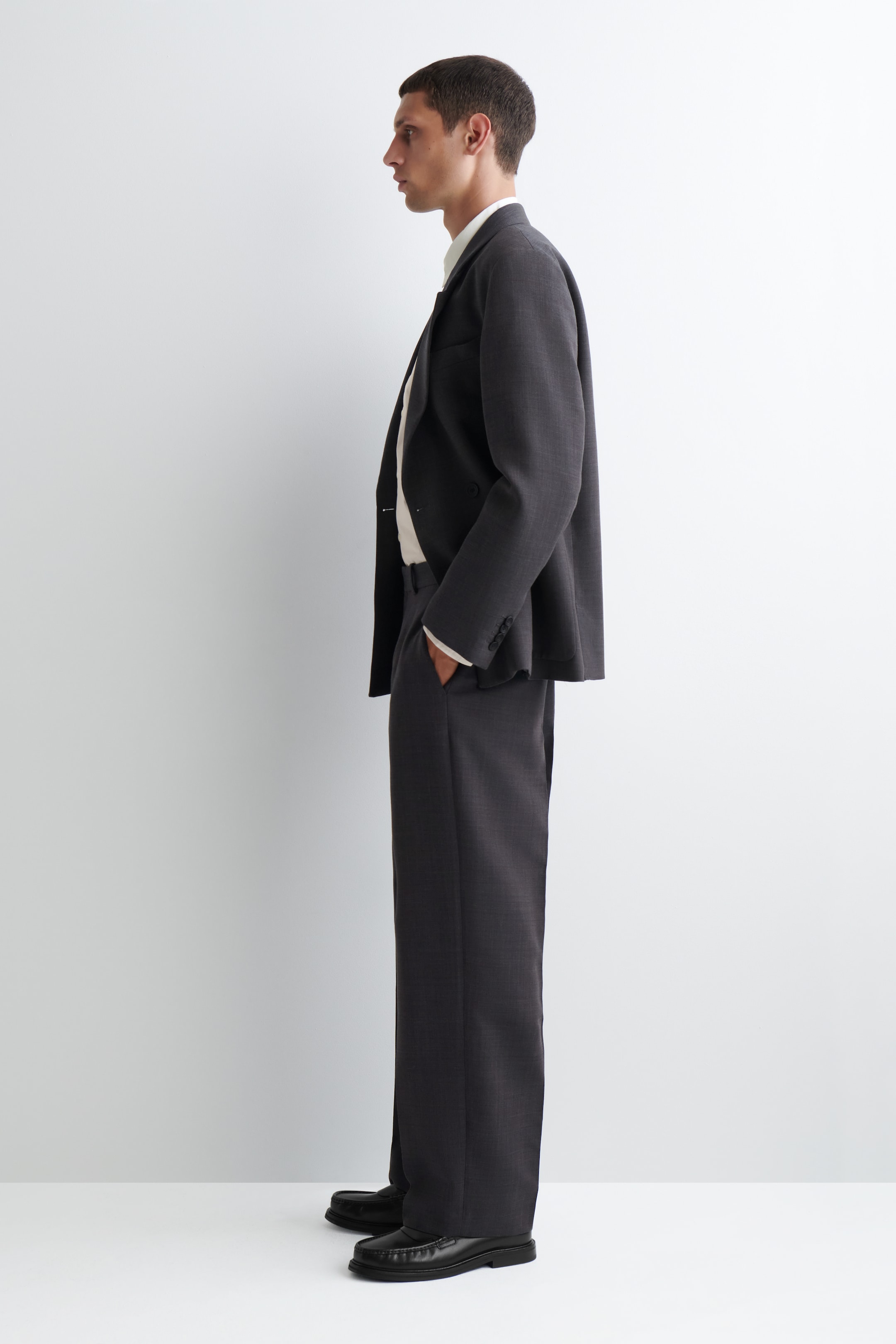 Agrandir l'image: Un costume gris foncé est présenté de profil, avec une veste croisée à coupe décontractée dotée de trois boutons de manchette visibles et d'un pantalon à jambes larges. La veste est portée sur une chemise de couleur claire, et le pantalon est assorti de mocassins noirs.