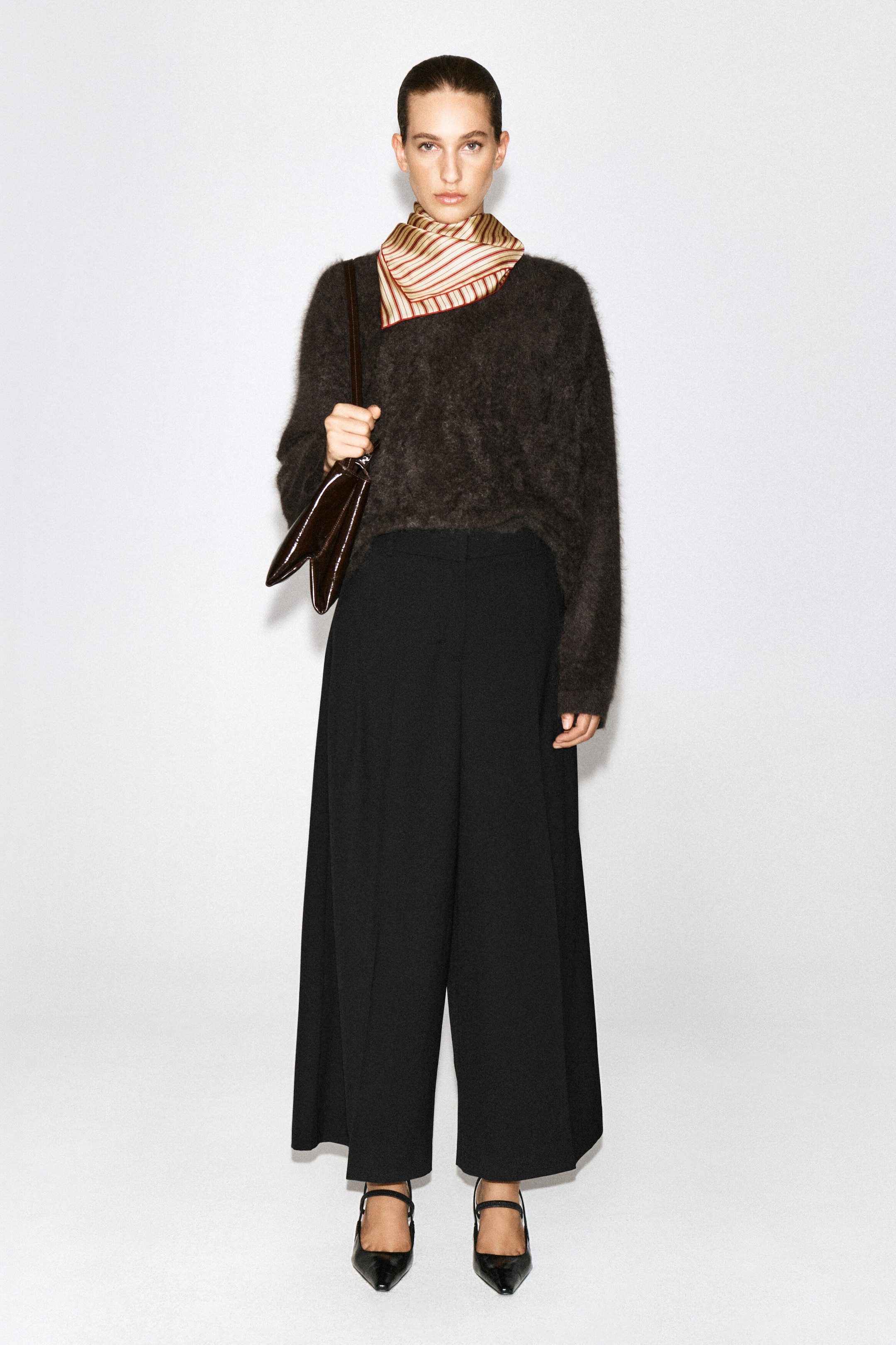 Pleated wool-blend wide-leg trousers in black are being worn by the model, featuring a relaxed wide-leg silhouette with front creases. The trousers are styled with a dark brown textured knit jumper, a striped neck scarf, and black pointed-toe heels. Pleated wool-blend wide-leg trousers in black are being worn by the model, featuring a relaxed wide-leg silhouette with front creases. The trousers are styled with a dark brown textured knit jumper, a striped neck scarf, and black pointed-toe heels.