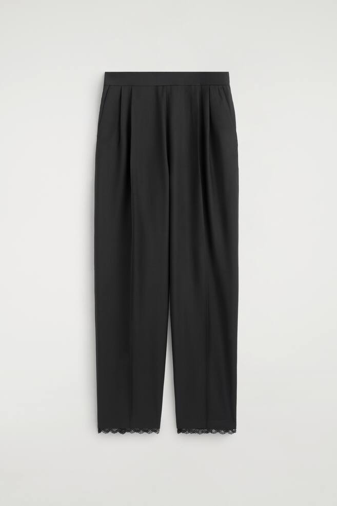 A pair of black trousers is hanging facing forwards, featuring a high-waisted design with deep front pleats and a relaxed, straight-leg silhouette. The trousers display a subtle front crease down each leg and are finished with a delicate black lace trim along the bottom hems. - 1