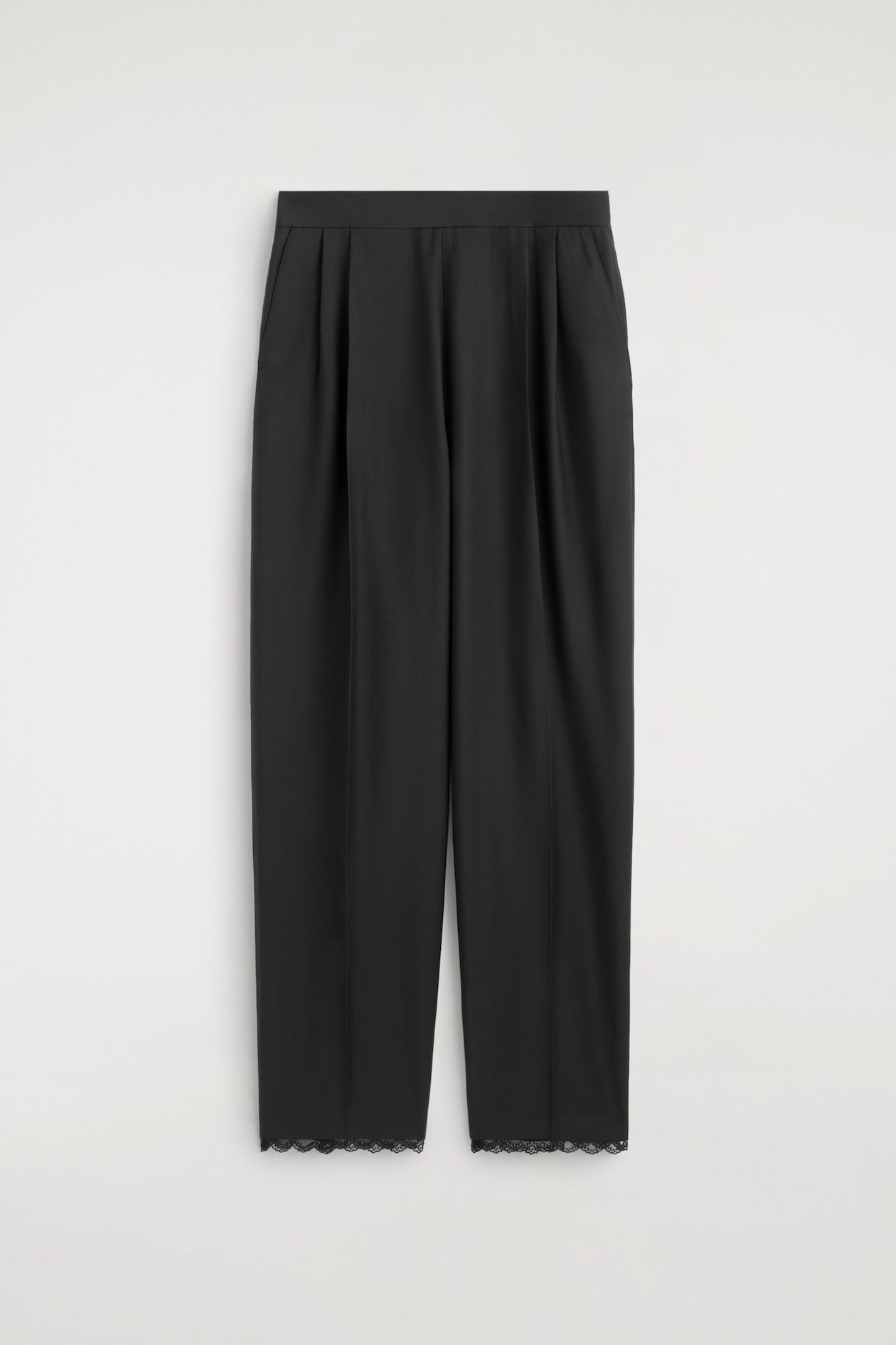 A pair of black trousers hangs facing forward, featuring a high-waisted design with deep front pleats and a relaxed, straight-leg silhouette. The trousers display a subtle front crease down each leg and finish with a delicate black lace trim along the bottom hems.
