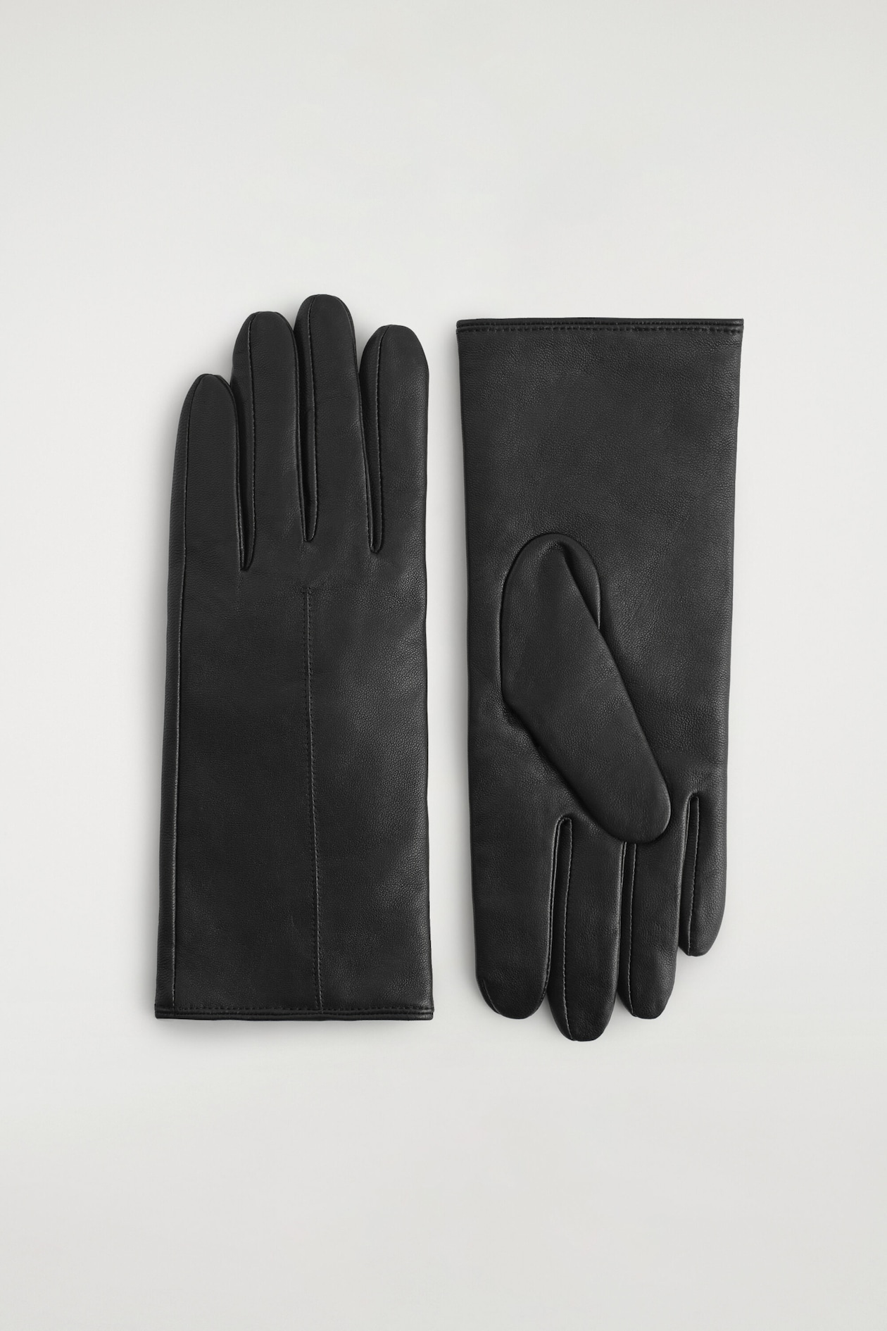 Une paire de gants en cuir noir est posée à plat côte à côte, montrant les détails de l'avant et de l'arrière du design. Le gant de gauche présente le dos de la main lisse avec une seule couture verticale piquée, tandis que le gant de droite affiche la paume avec un pouce replié et des ourlets cousus nets.