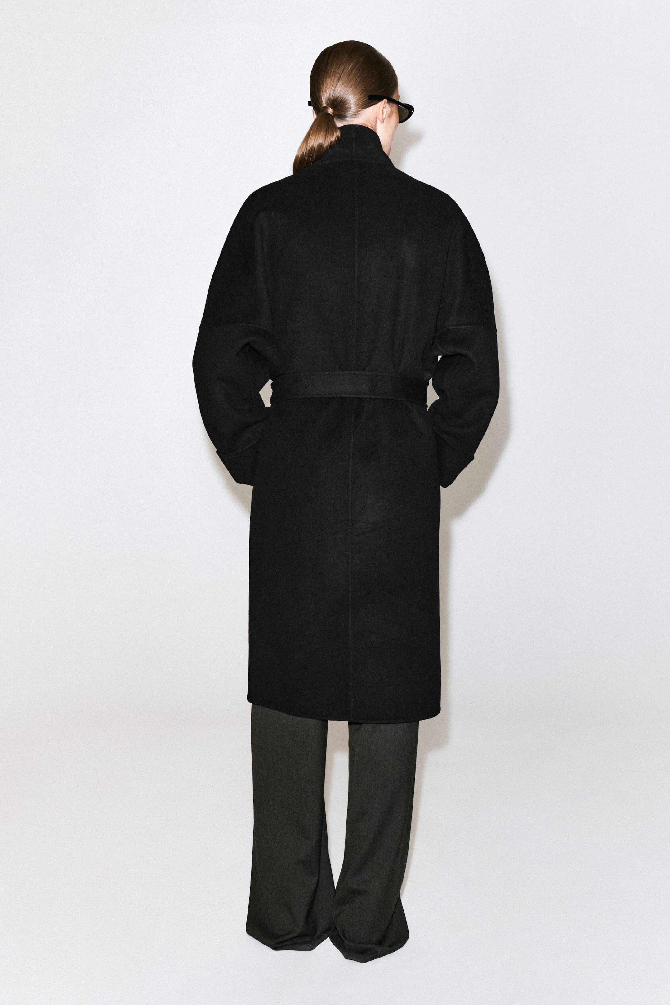 Un manteau châle oversize en laine double face noir est porté par le mannequin, vu de dos. Ce manteau mi-long présente des épaules tombantes et une ceinture assortie en tissu nouée à la taille, porté sur un pantalon sombre à jambes larges. Un manteau châle oversize en laine double face noir est porté par le mannequin, vu de dos. Ce manteau mi-long présente des épaules tombantes et une ceinture assortie en tissu nouée à la taille, porté sur un pantalon sombre à jambes larges.