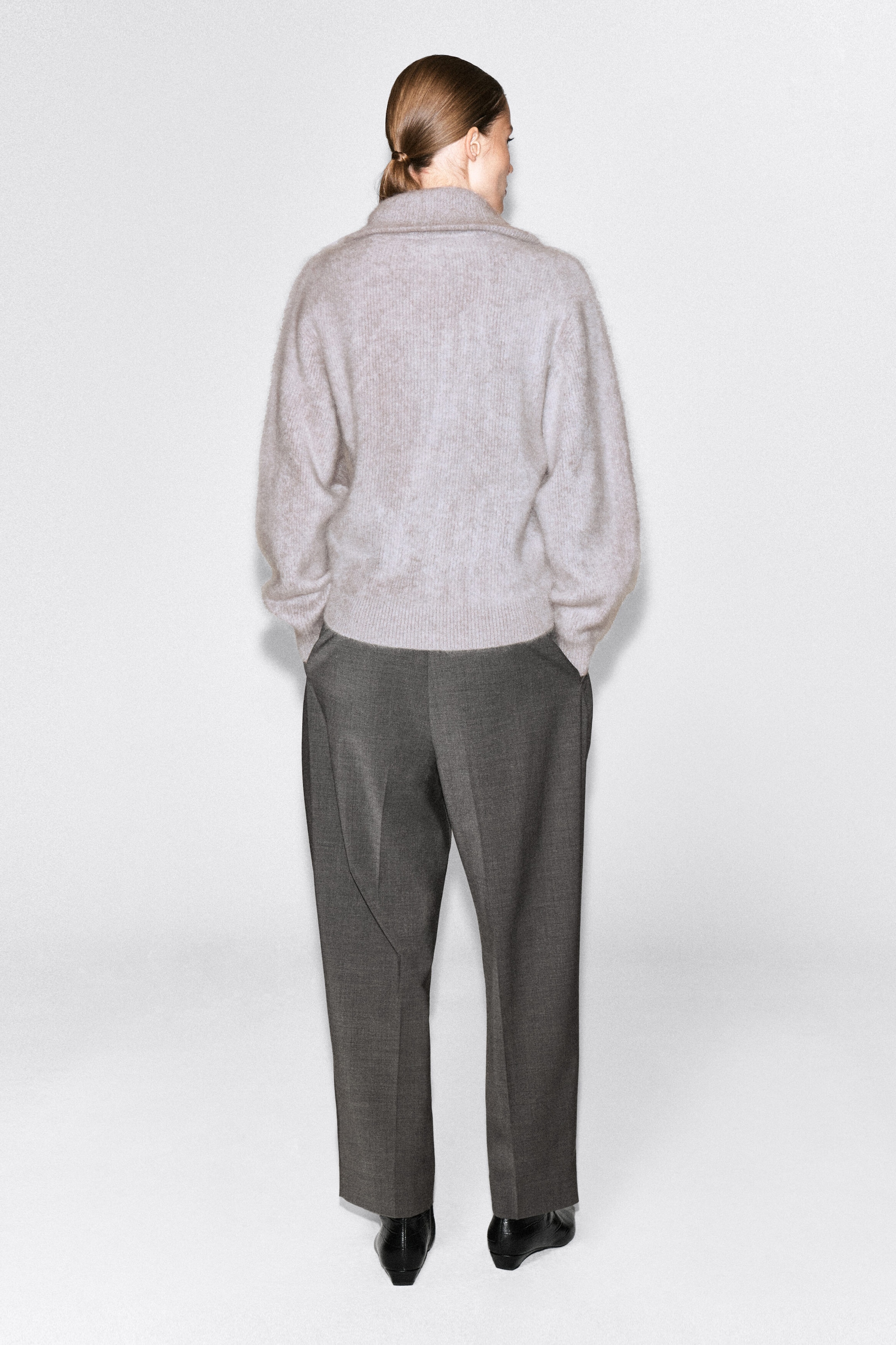 The model is wearing a light gray brushed cashmere sweater, standing with their back to the camera. The sweater features a high neckline and ribbed hem and is paired with dark gray trousers. The model is wearing a light gray brushed cashmere sweater, standing with their back to the camera. The sweater features a high neckline and ribbed hem and is paired with dark gray trousers.