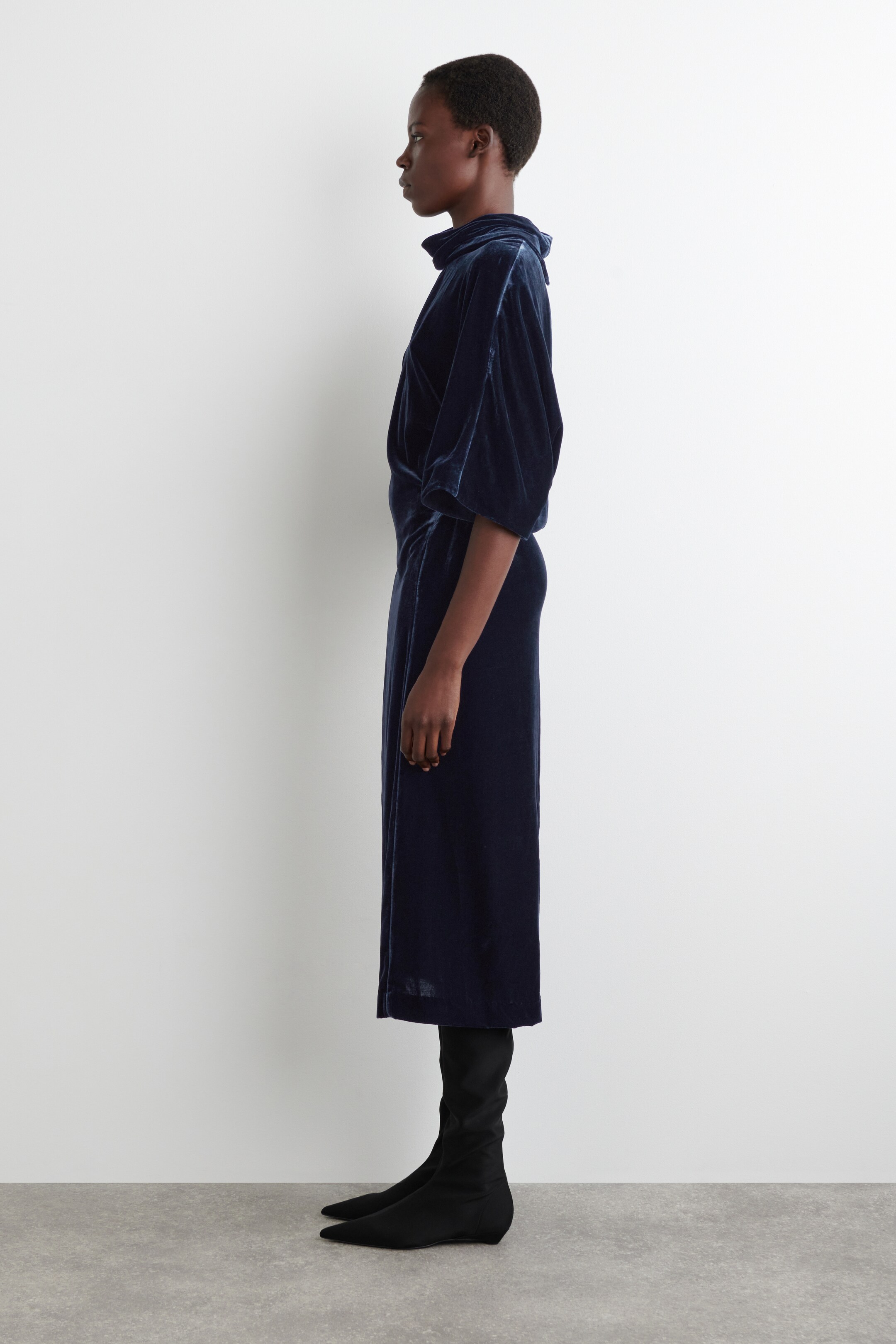 Ver imagem maior: O vestido de veludo azul-marinho é apresentado de perfil lateral, virado para a esquerda, com um decote alto tipo cowl e mangas volumosas. As mangas estão reunidas numa cintura ajustada, e o vestido estende-se numa saia de comprimento midi.
