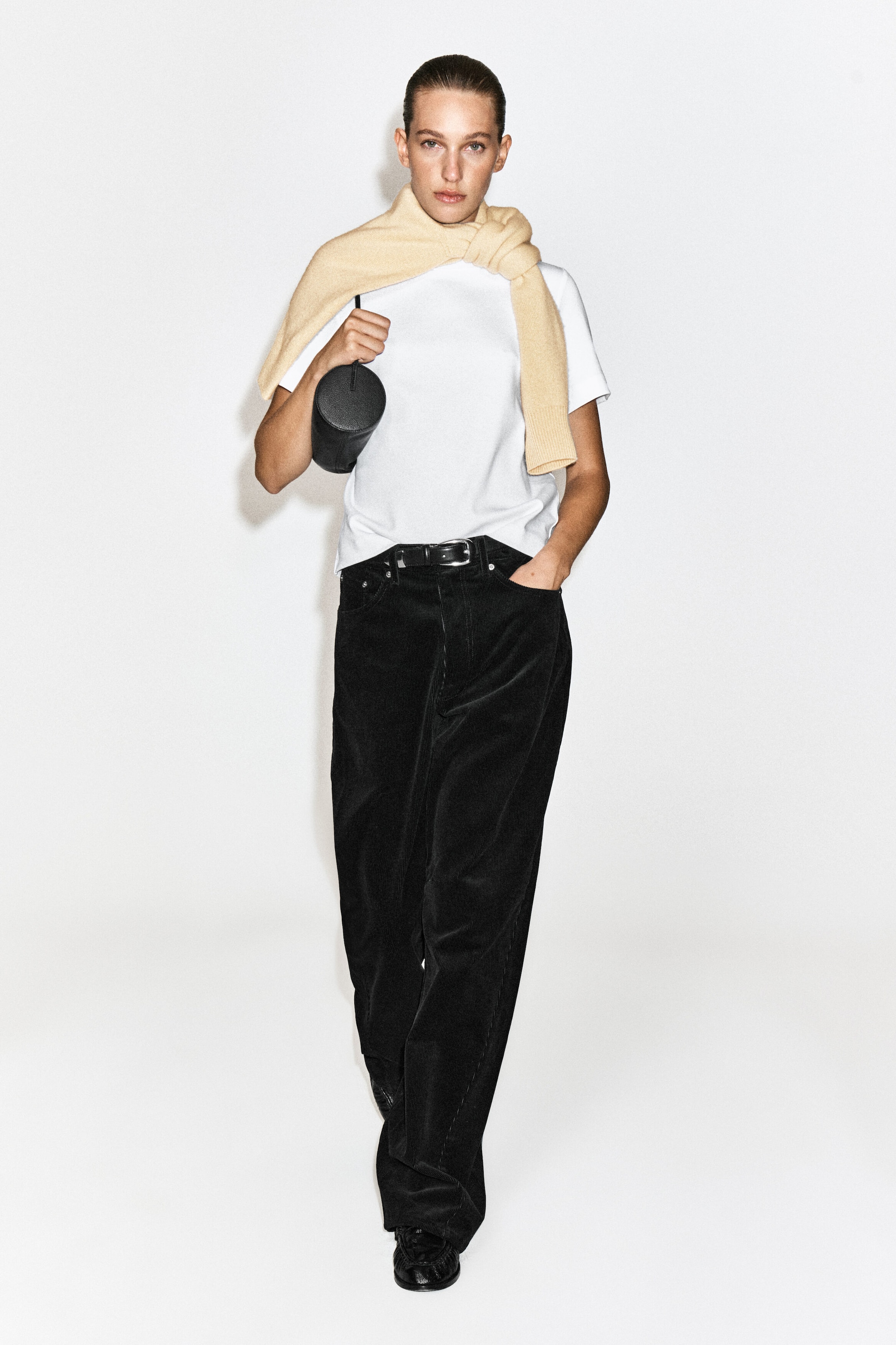 The model is wearing black wide-leg corduroy trousers, styled with a black leather belt, a white T-shirt, and a light beige jumper draped over the shoulders. The model is also carrying a small, cylindrical black leather bag in one hand.
