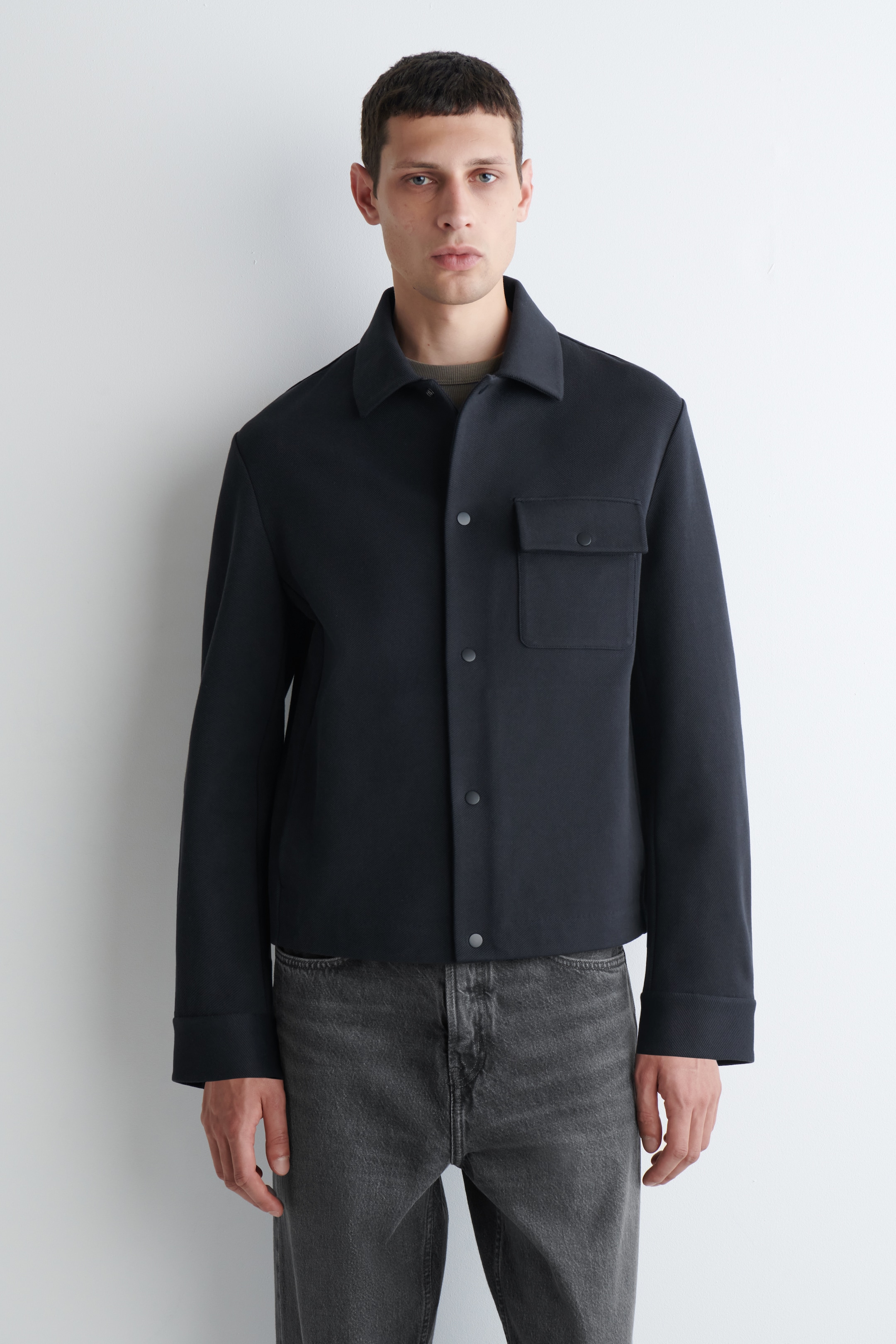 Agrandir l'image: Une veste bleu foncé est portée de face, avec un col pointu et une fermeture à pression visible. Elle présente une seule poche plaquée à rabat sur la poitrine gauche et des manches longues avec des poignets simples, se terminant par un ourlet droit à la taille. Un haut vert foncé est superposé en dessous.