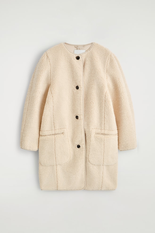 Eine cremefarbene Longline-Teddyjacke wird von vorne präsentiert. Sie verfügt über einen abgerundeten, kragenlosen Ausschnitt, einen Knopfverschluss mit dunklen Knöpfen, zwei große aufgesetzte Taschen und paspelierte Kanten. - 1