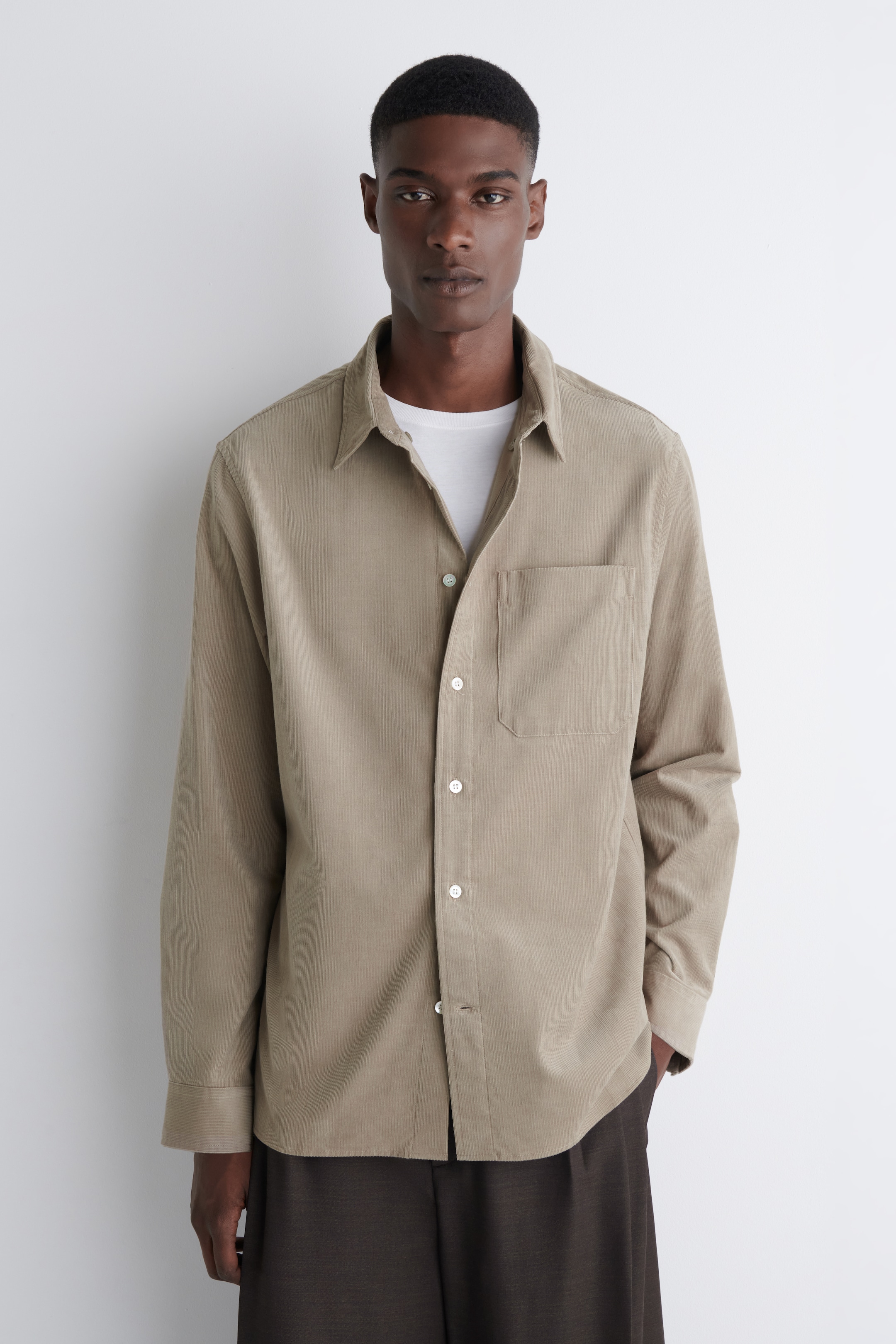 View larger image: A beige corduroy shirt is being presented from the front, featuring a pointed collar, long sleeves with buttoned cuffs, and a patch pocket on the left chest. It is being buttoned down the front with white buttons and is being layered over a white crew-neck t-shirt.