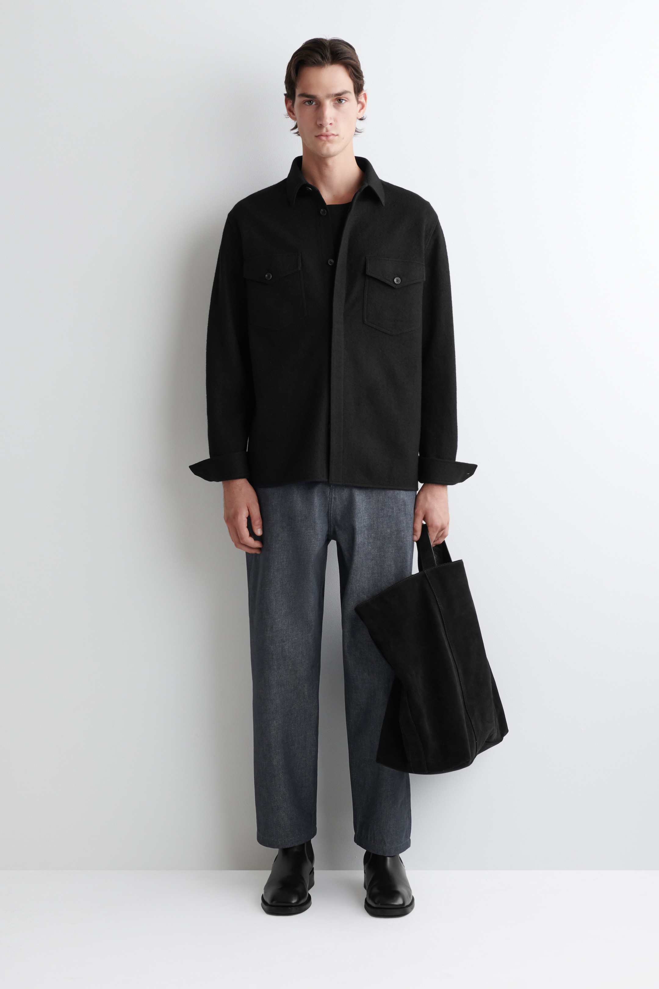 Agrandir l'image: L'overshirt noir apparaît de face, avec une coupe décontractée dans un tissu doux et texturé. Il est conçu avec un col pointu et deux poches à rabat boutonnées sur la poitrine, tandis que ses manches longues sont retroussées aux poignets et il est superposé sur un t-shirt à col rond foncé.