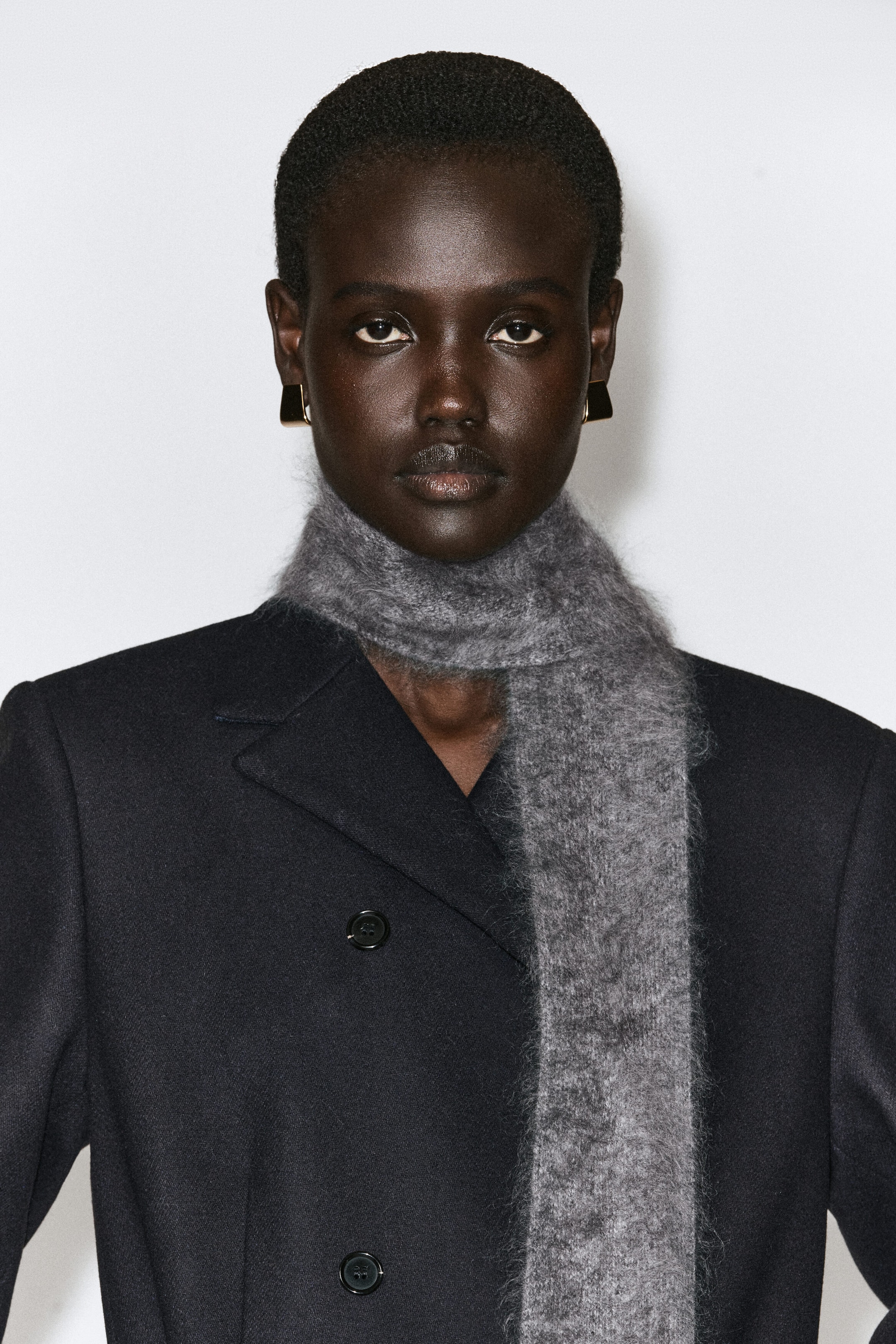 A narrow, brushed gray wool scarf is shown from the front, wrapped once around the neck with one end hanging down the front of a dark double-breasted coat. The scarf showcases a highly tactile, fuzzy texture and a slim, tailored silhouette. A narrow, brushed gray wool scarf is shown from the front, wrapped once around the neck with one end hanging down the front of a dark double-breasted coat. The scarf showcases a highly tactile, fuzzy texture and a slim, tailored silhouette.