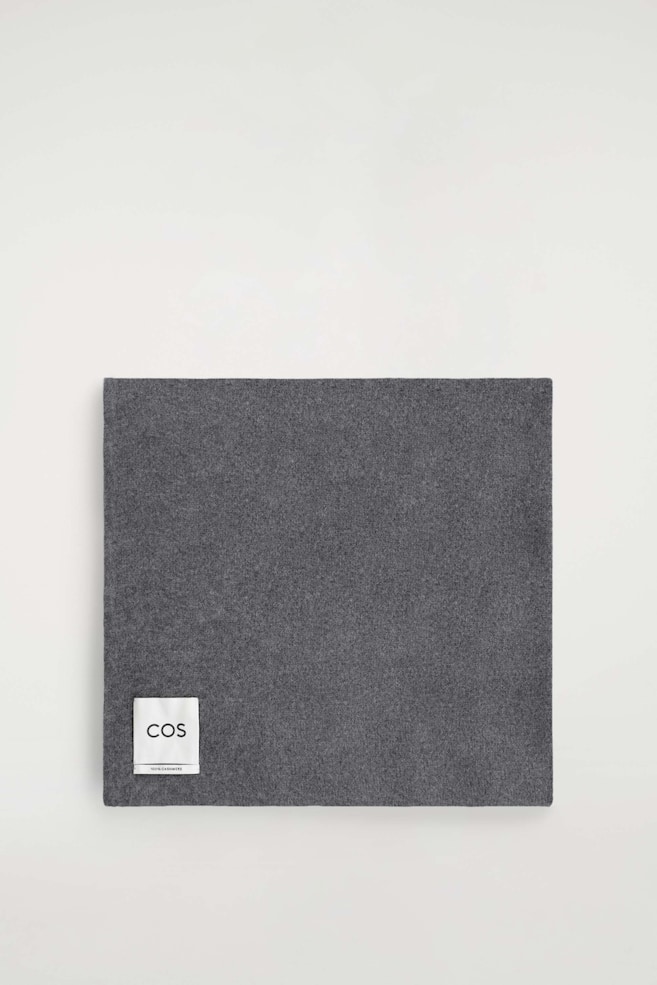 A cashmere scarf is laid flat and folded into a square, showcasing its dark grey melange colour and soft, fine-knit texture. A white brand label is positioned in the bottom-left corner, displaying the "COS" logo and "100% CASHMERE" text. - 1