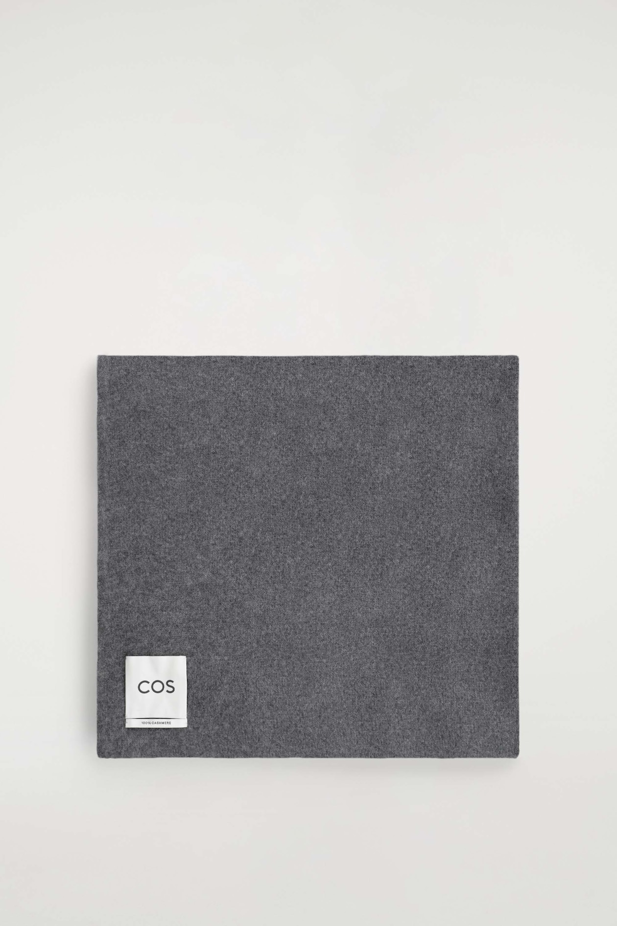 A cashmere scarf is laid flat and folded into a square, showcasing its dark grey melange colour and soft, fine-knit texture. A white brand label is positioned in the bottom-left corner, displaying the "COS" logo and "100% CASHMERE" text.