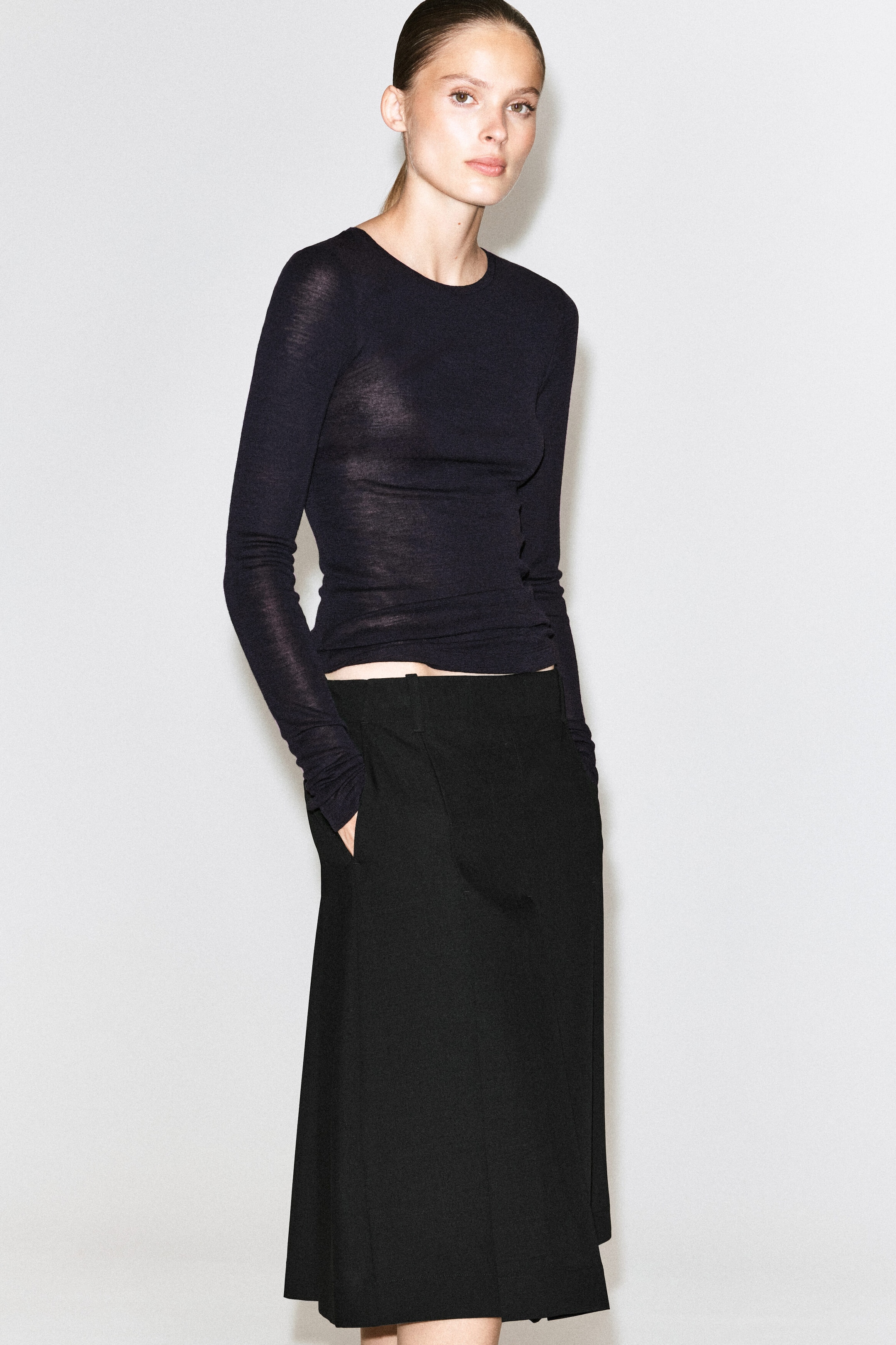 A black box-pleat wool midi skirt is being worn by the model, who is standing with one hand placed in the side pocket. The structured skirt is styled with a fitted, long-sleeve black top. A black box-pleat wool midi skirt is being worn by the model, who is standing with one hand placed in the side pocket. The structured skirt is styled with a fitted, long-sleeve black top.