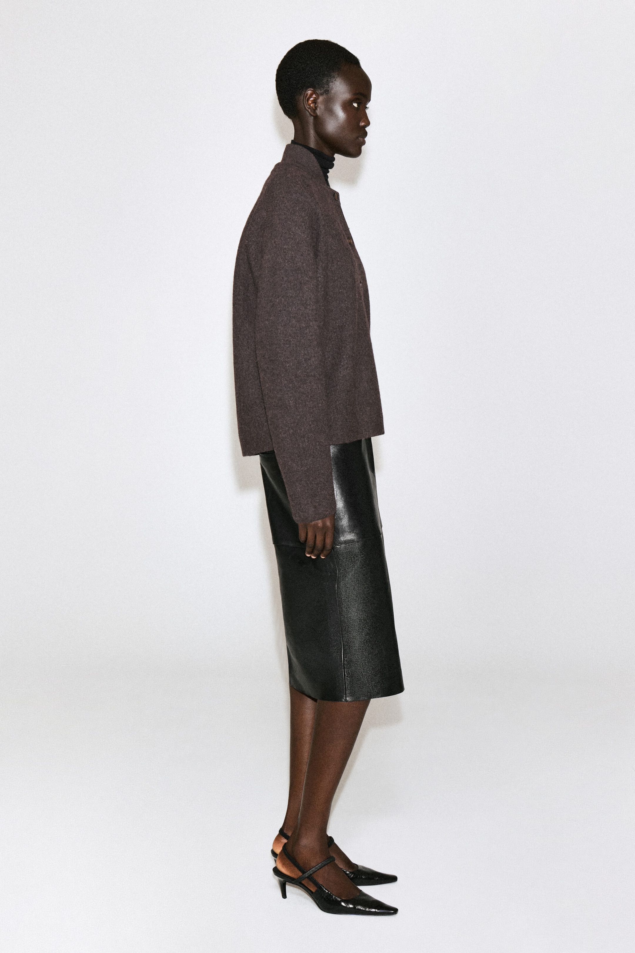 Un cardigan en laine bouillie brun foncé est porté de profil, face vers la droite, mettant en valeur sa silhouette doucement arrondie et son col montant superposé sur un col roulé noir. Le cardigan est agencé avec une jupe midi en cuir noir et des escarpins noirs à bout pointu afin de mettre en valeur sa structure et sa coupe. Un cardigan en laine bouillie brun foncé est porté de profil, face vers la droite, mettant en valeur sa silhouette doucement arrondie et son col montant superposé sur un col roulé noir. Le cardigan est agencé avec une jupe midi en cuir noir et des escarpins noirs à bout pointu afin de mettre en valeur sa structure et sa coupe.