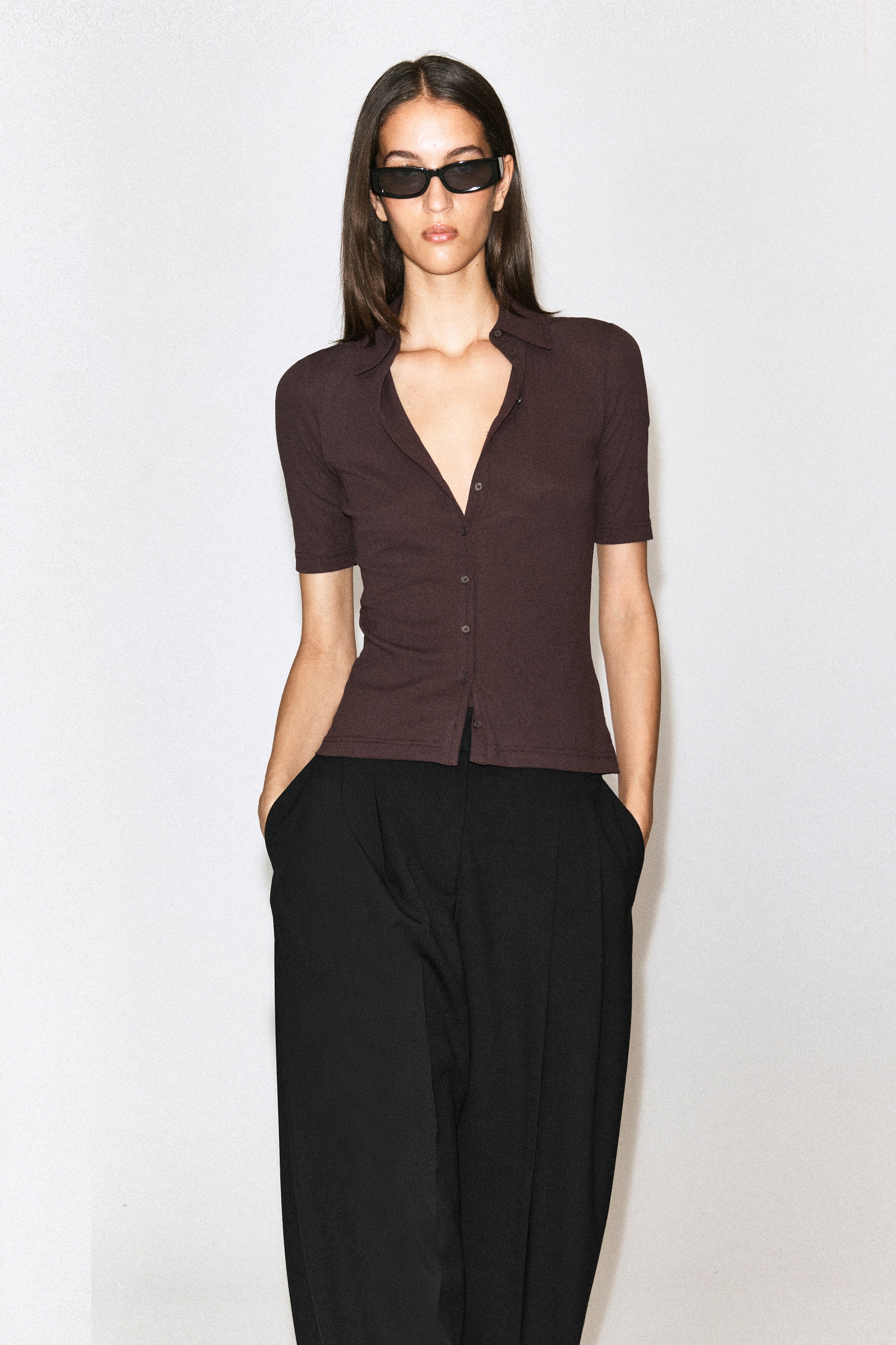 The model is wearing a dark brown, short-sleeved cotton jersey shirt with a classic collar and button-front closure. The slim-fit shirt is styled partially unbuttoned at the neckline and paired with high-waisted, wide-leg black trousers. The model is wearing a dark brown, short-sleeved cotton jersey shirt with a classic collar and button-front closure. The slim-fit shirt is styled partially unbuttoned at the neckline and paired with high-waisted, wide-leg black trousers.