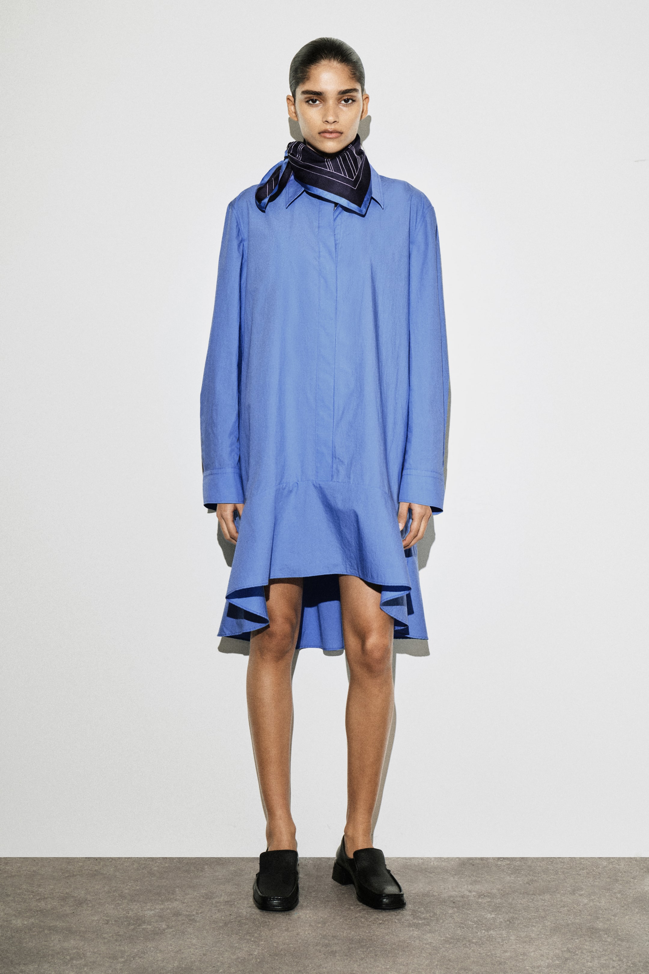 Ver imagen más grande: Un vestido camisero azul cielo se ve desde el frente, con un cuello alargado en punta y una tapeta oculta. Está diseñado con una cintura caída y un dobladillo acampanado que es visiblemente más largo en la parte trasera.