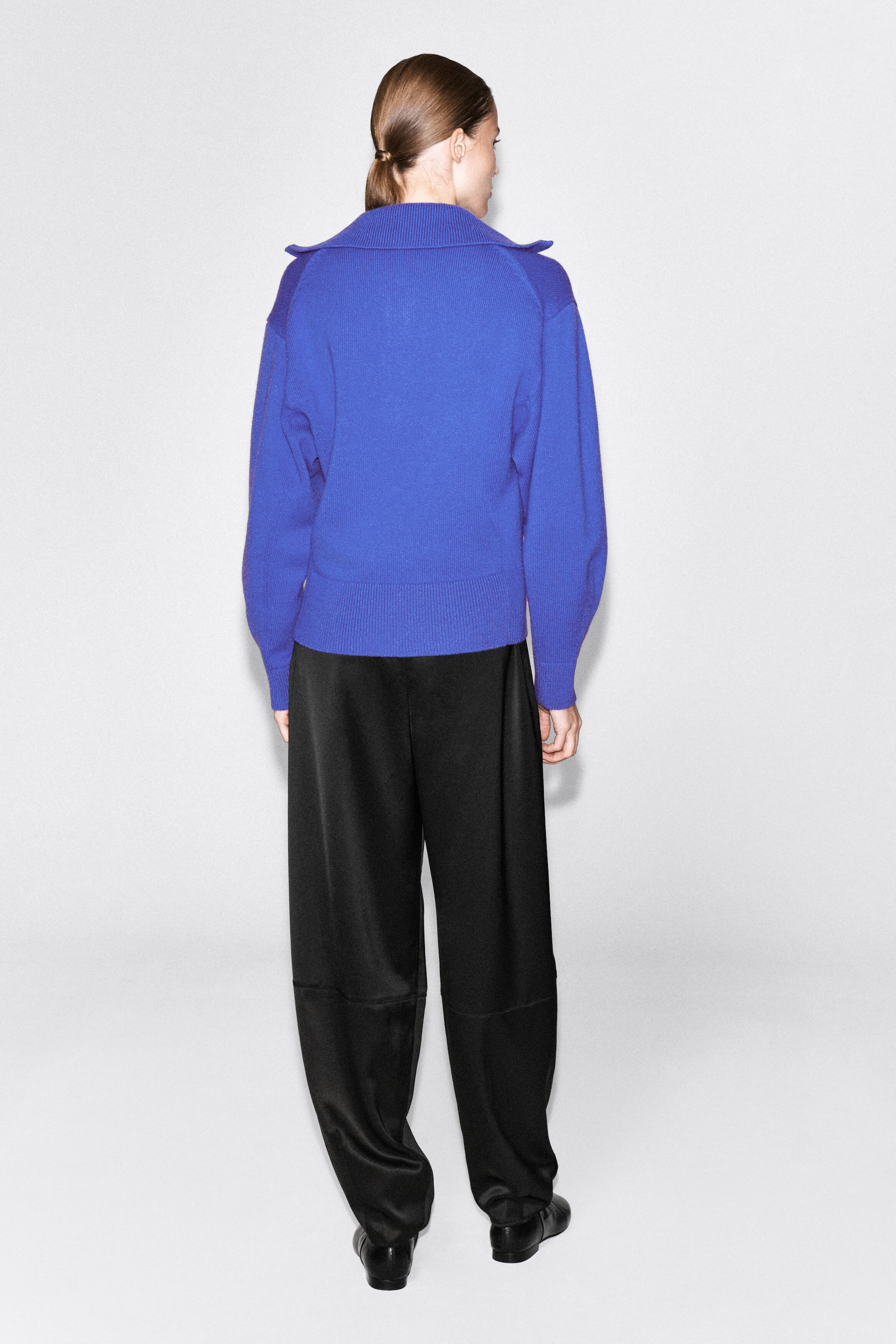 A bright blue knit sweater featuring a wide ribbed collar, relaxed sleeves, and a ribbed hem is being worn by the model, shown from the back. The sweater is paired with black wide-leg trousers. A bright blue knit sweater featuring a wide ribbed collar, relaxed sleeves, and a ribbed hem is being worn by the model, shown from the back. The sweater is paired with black wide-leg trousers.