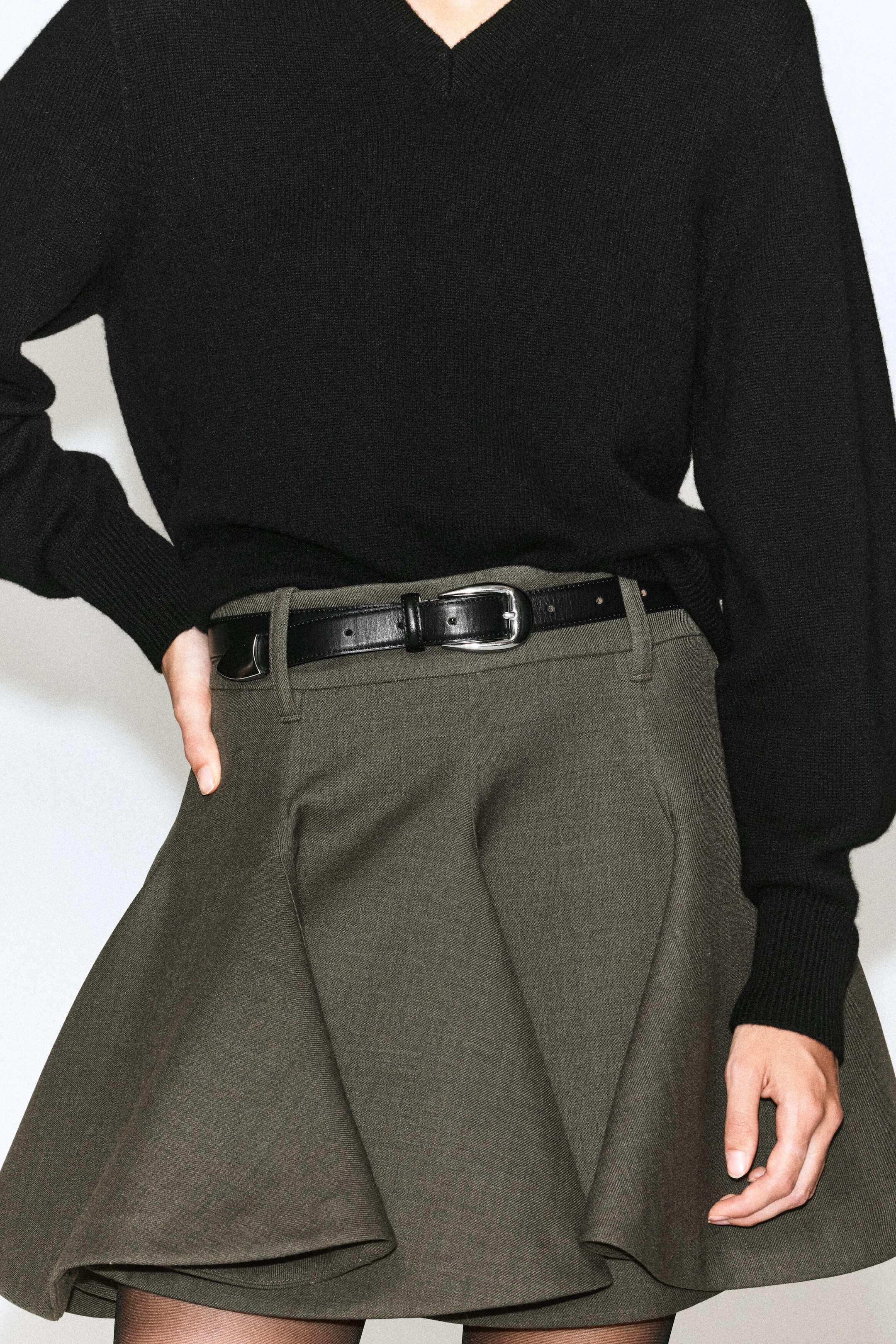 A sculptural tailored wool mini skirt in olive green is worn by the model, styled with a black leather belt and a tucked-in black V-neck sweater. The skirt features a structured, flared silhouette with deep pleats, and the model rests one hand near the waistband. A sculptural tailored wool mini skirt in olive green is worn by the model, styled with a black leather belt and a tucked-in black V-neck sweater. The skirt features a structured, flared silhouette with deep pleats, and the model rests one hand near the waistband.