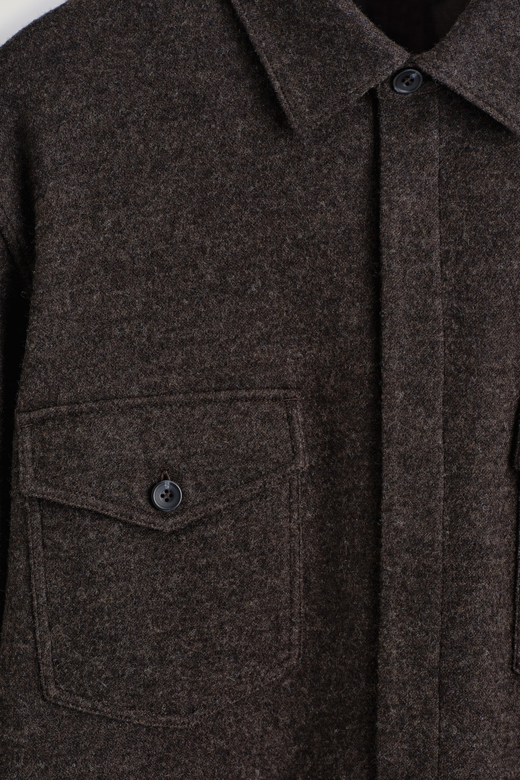 Agrandir l'image: Une surchemise brun foncé est présentée de face, mettant en valeur son tissu texturé en mélange de laine bouillie et de coton. Elle est dotée d'un col pointu, d'une patte de boutonnage dissimulée avec un bouton supérieur visible, et d'une poche plaquée avec un rabat boutonné sur la poitrine gauche, le tout accentué par des surpiqûres ton sur ton.