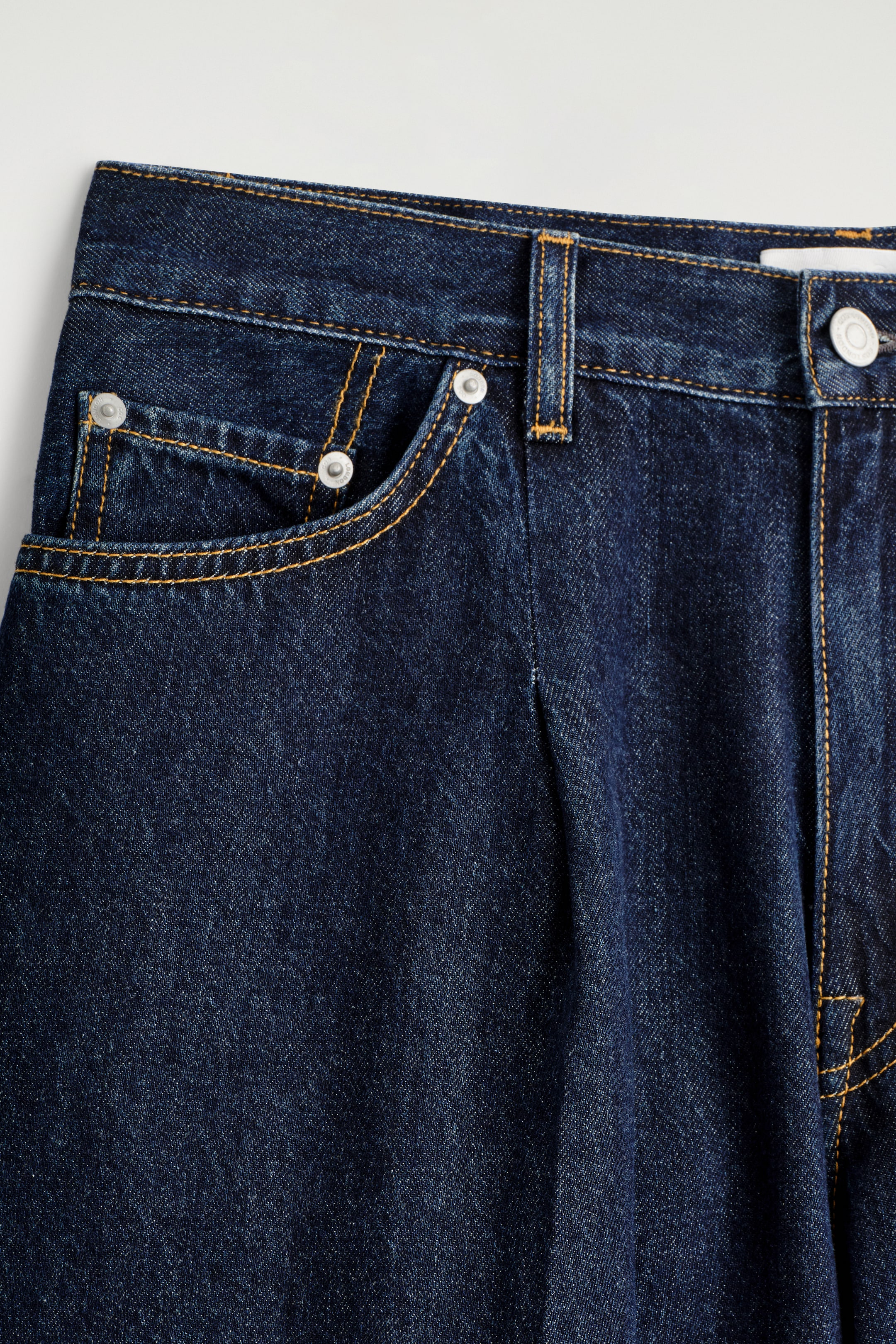 Visualizza immagine più grande: I jeans in denim indaco scuro presentano una tasca frontale curva con rivetti color argento, cuciture a contrasto giallo-arancio e una singola piega visibile sotto la cintura. Un bottone color argento fissa la cintura.