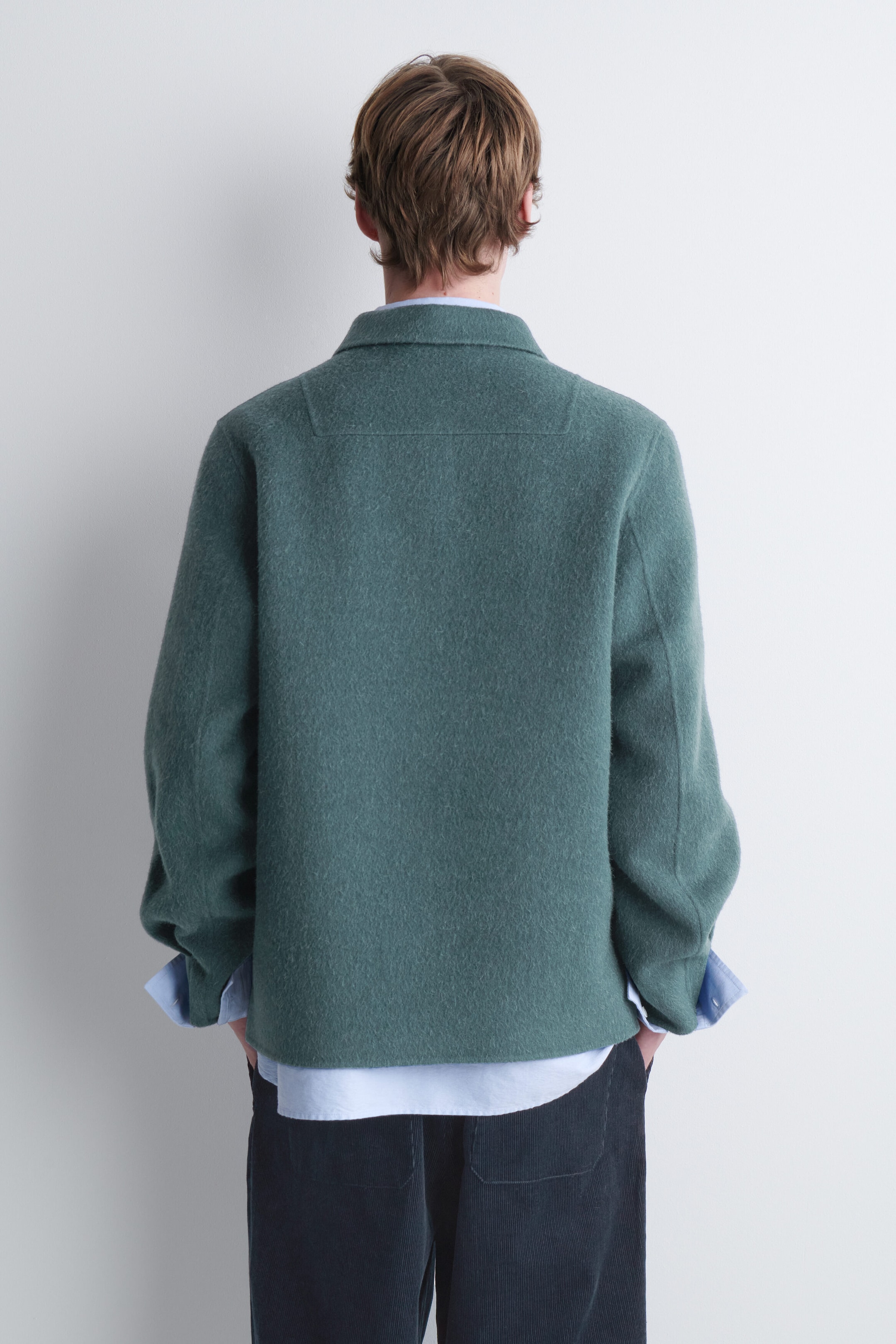 A green wool jacket with a textured finish is being worn by the model, shown from the back. The jacket features a classic collar, a straight hem, and a back yoke, and is layered over a light blue collared shirt and dark trousers. A green wool jacket with a textured finish is being worn by the model, shown from the back. The jacket features a classic collar, a straight hem, and a back yoke, and is layered over a light blue collared shirt and dark trousers.