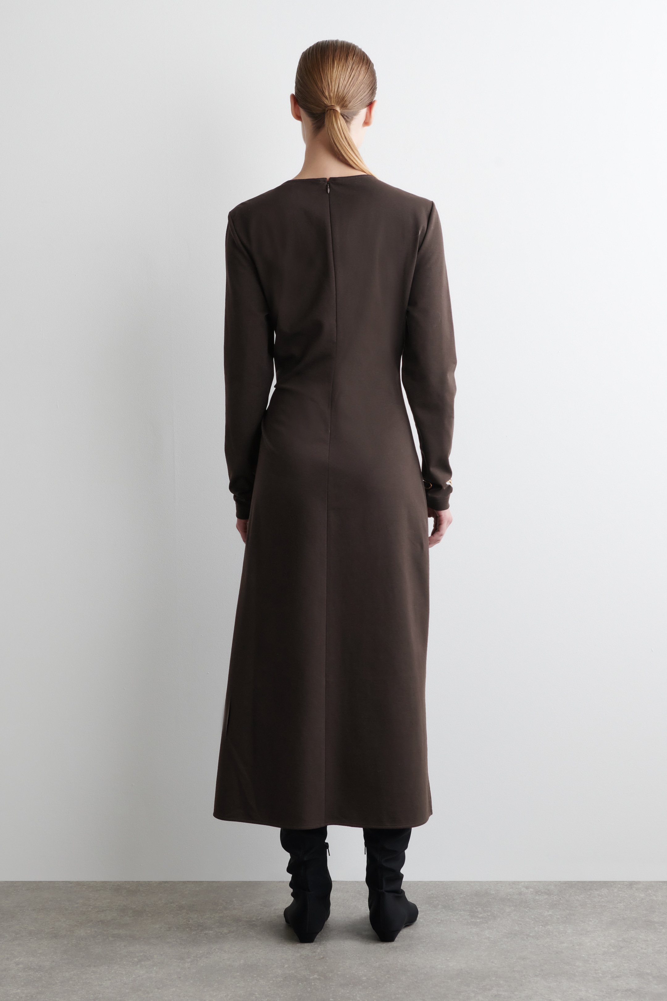 Agrandir l'image: Une robe maxi brun foncé est présentée de dos, avec des épaules structurées, des manches longues et un corsage ajusté. Une fermeture éclair se trouve au centre du dos, et la robe tombe jusqu'aux chevilles, couvrant des bottes noires.