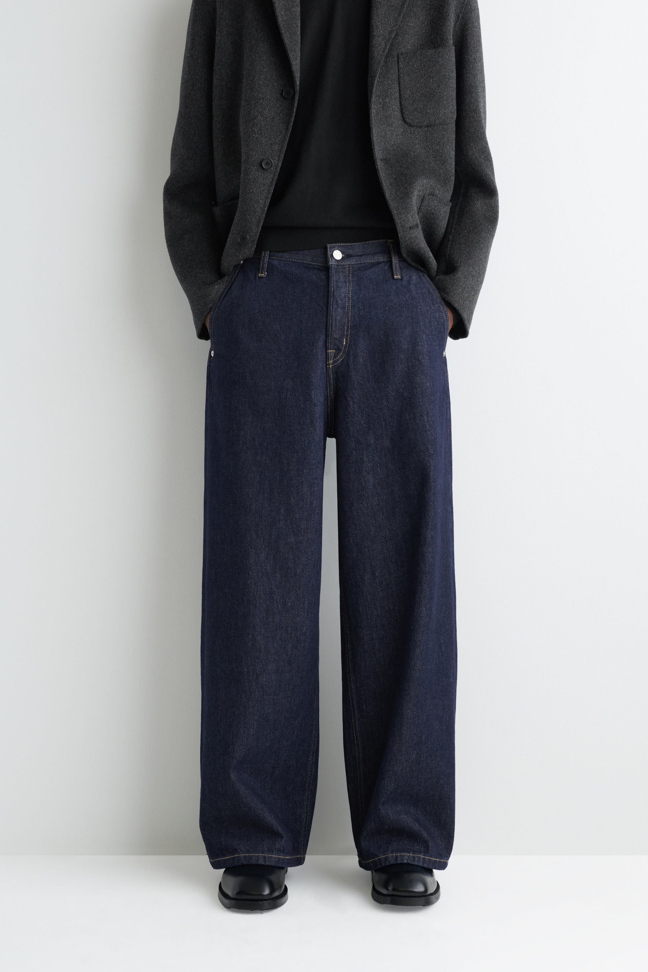 Agrandir l'image: Un jean en denim à jambes larges et lavage foncé est présenté de face, avec des coutures contrastées visibles, des passants de ceinture et une fermeture à bouton unique à la taille. Le jean présente une coupe décontractée et pleine longueur, avec l'ourlet reposant sur le dessus des chaussures noires du porteur.