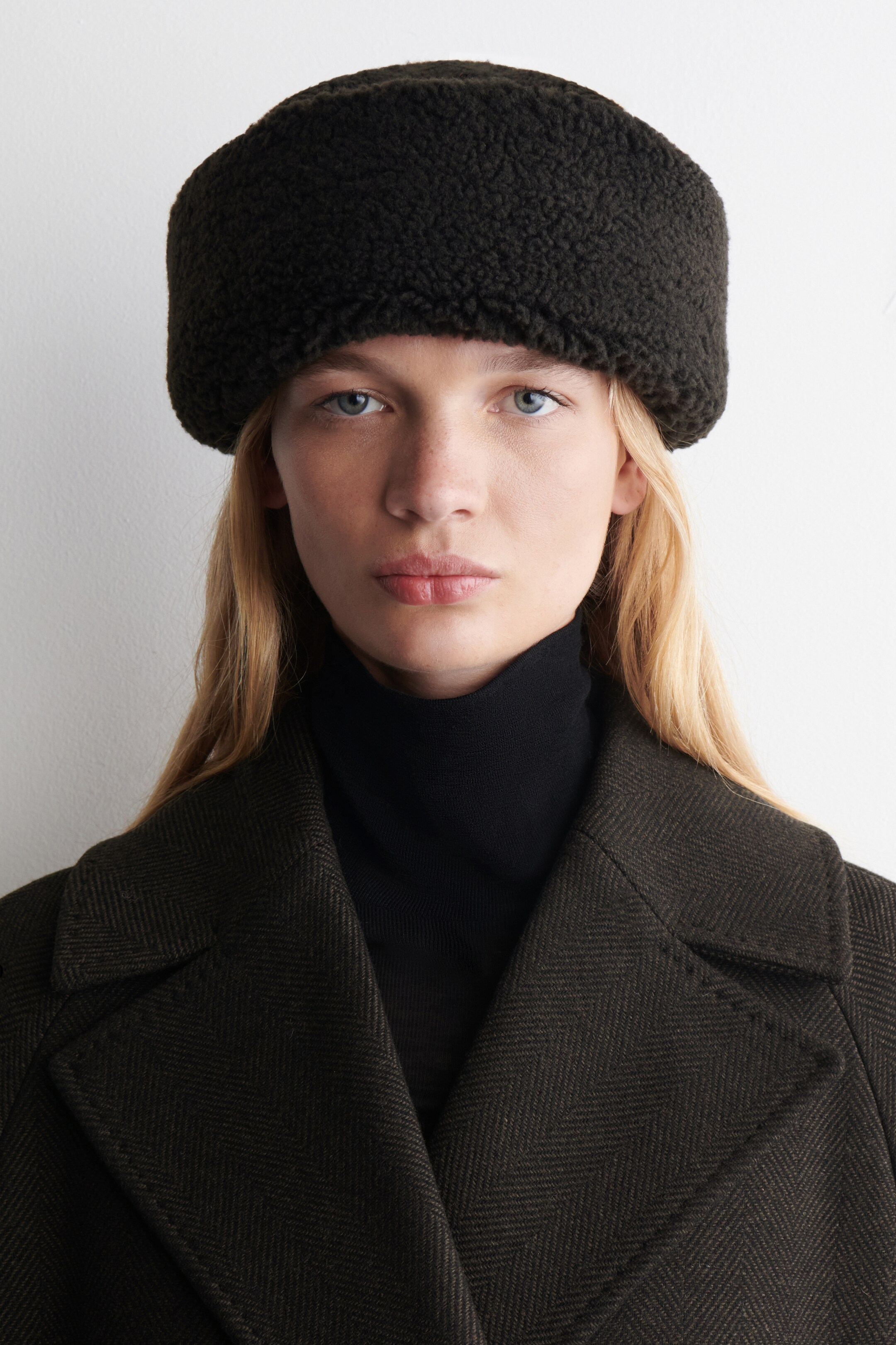 View larger image: A dark brown hat, made of a curly pile fabric, is covering the forehead, top of the head, and ears. A black turtleneck is layering under a dark brown herringbone coat featuring wide notch lapels.