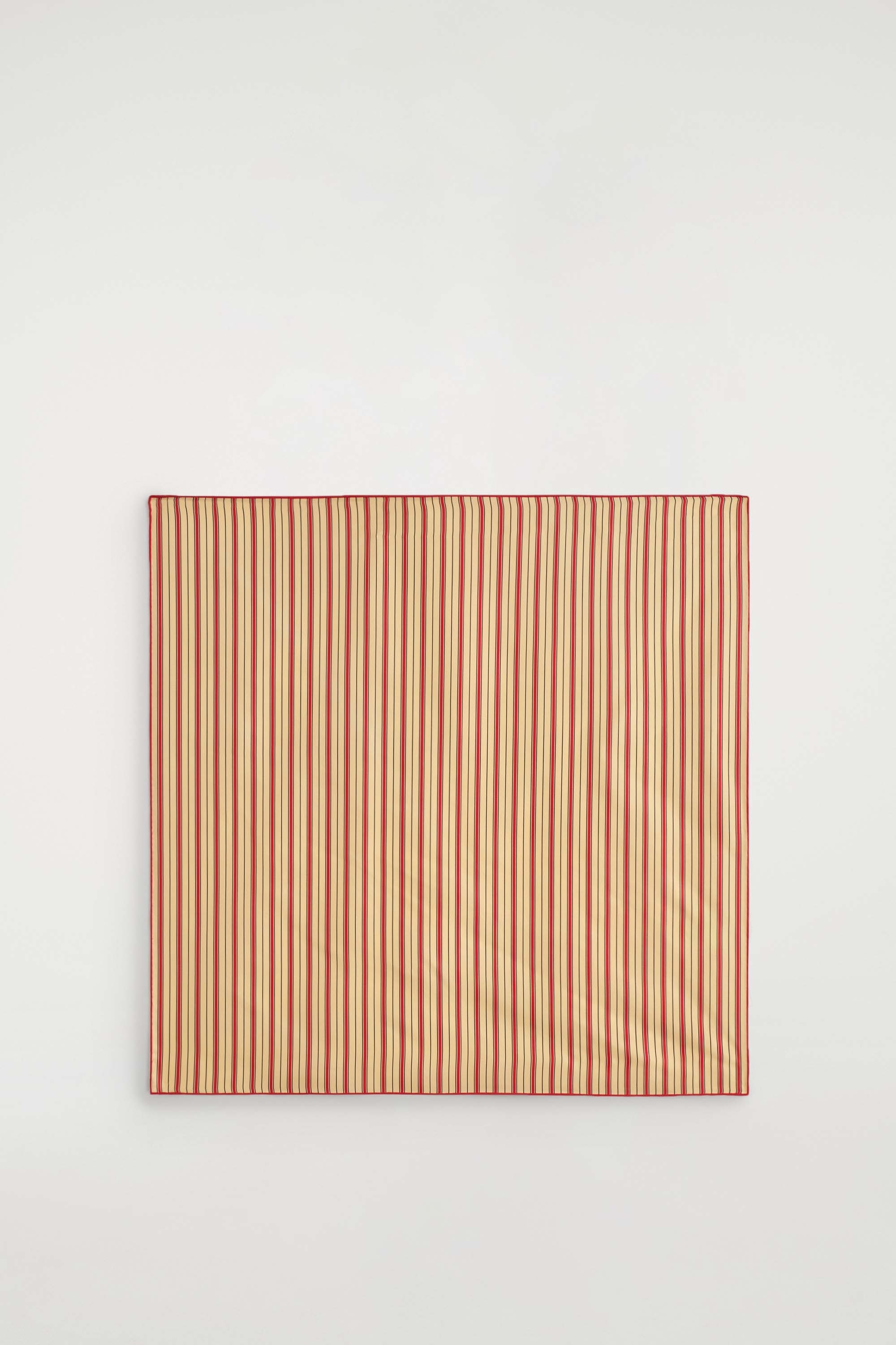 Une écharpe en soie à rayures, avec des rayures rouges et noires sur fond beige, est portée nouée autour du cou du mannequin. Lâécharpe est associée à un pull en maille de couleur crème.
