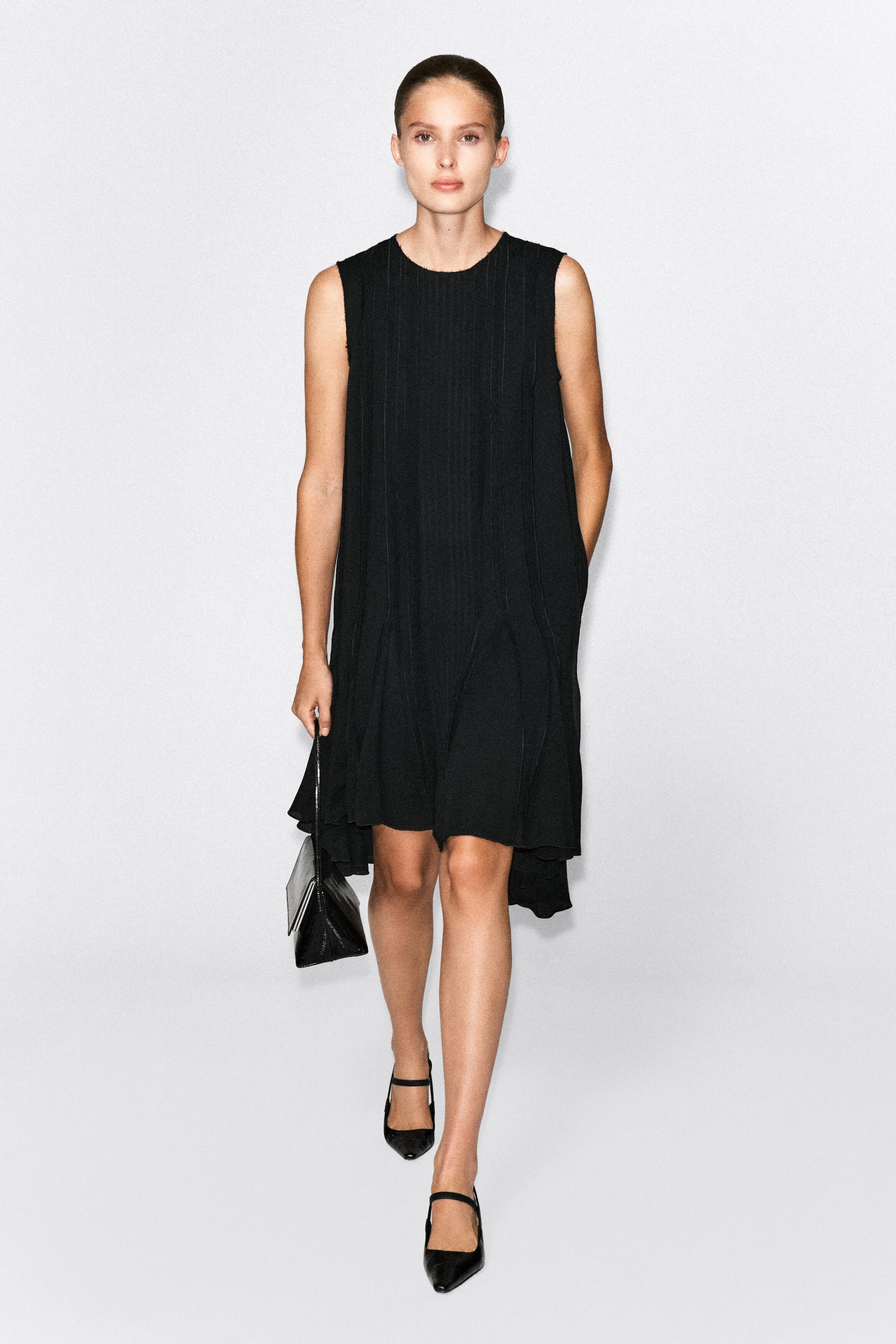 A voluminous black pleated midi dress, featuring a sleeveless design and a round neck, is worn by the model. The dress features vertical pleat detailing and a flared, asymmetric hemline, styled with a small black handbag and black pointed-toe flat shoes. A voluminous black pleated midi dress, featuring a sleeveless design and a round neck, is worn by the model. The dress features vertical pleat detailing and a flared, asymmetric hemline, styled with a small black handbag and black pointed-toe flat shoes.