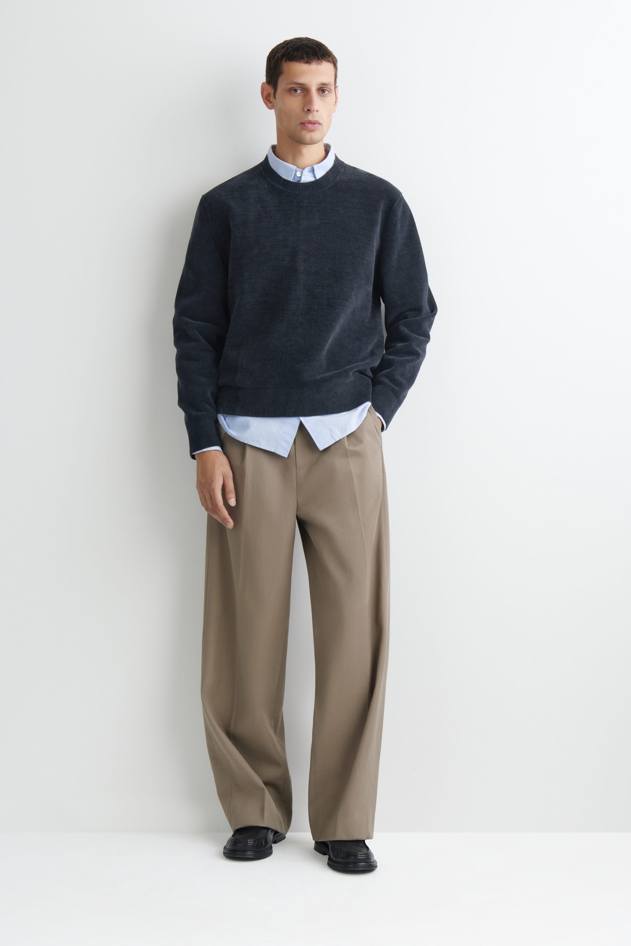 The dark gray cotton-chenille crewneck sweater is shown from the front, featuring a soft, textured finish with a ribbed collar, cuffs, and hem. It is layered over a light blue collared shirt, with the collar and untucked hem visible, and is styled with wide-leg beige trousers. The dark gray cotton-chenille crewneck sweater is shown from the front, featuring a soft, textured finish with a ribbed collar, cuffs, and hem. It is layered over a light blue collared shirt, with the collar and untucked hem visible, and is styled with wide-leg beige trousers.