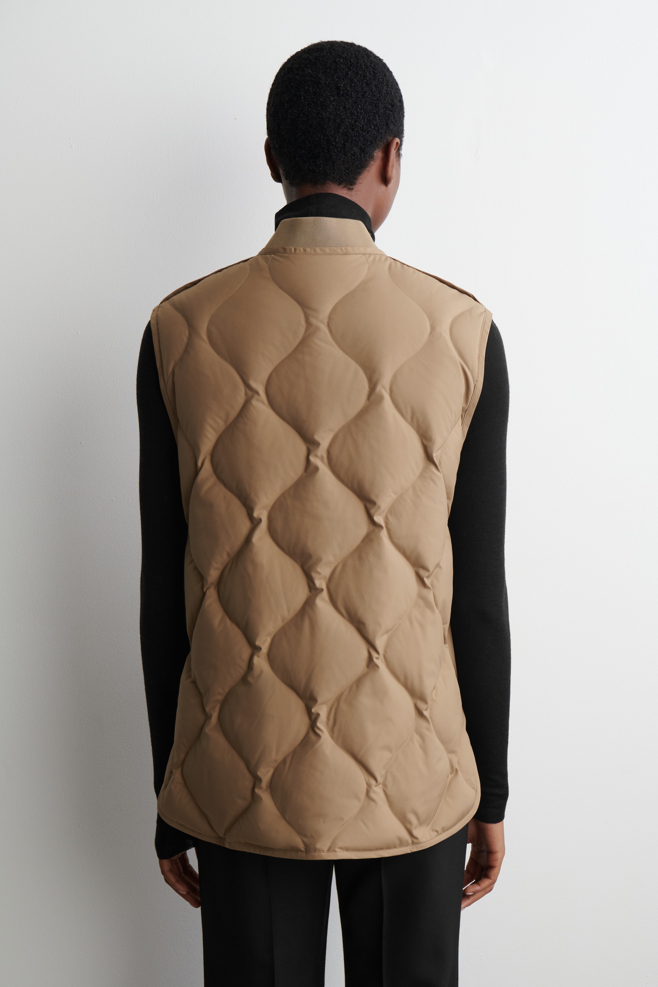 View larger image: A camel-colored quilted vest is being presented from the back, featuring an onion quilting pattern and a contrasting light beige stand collar. It is being worn over a black long-sleeved top.