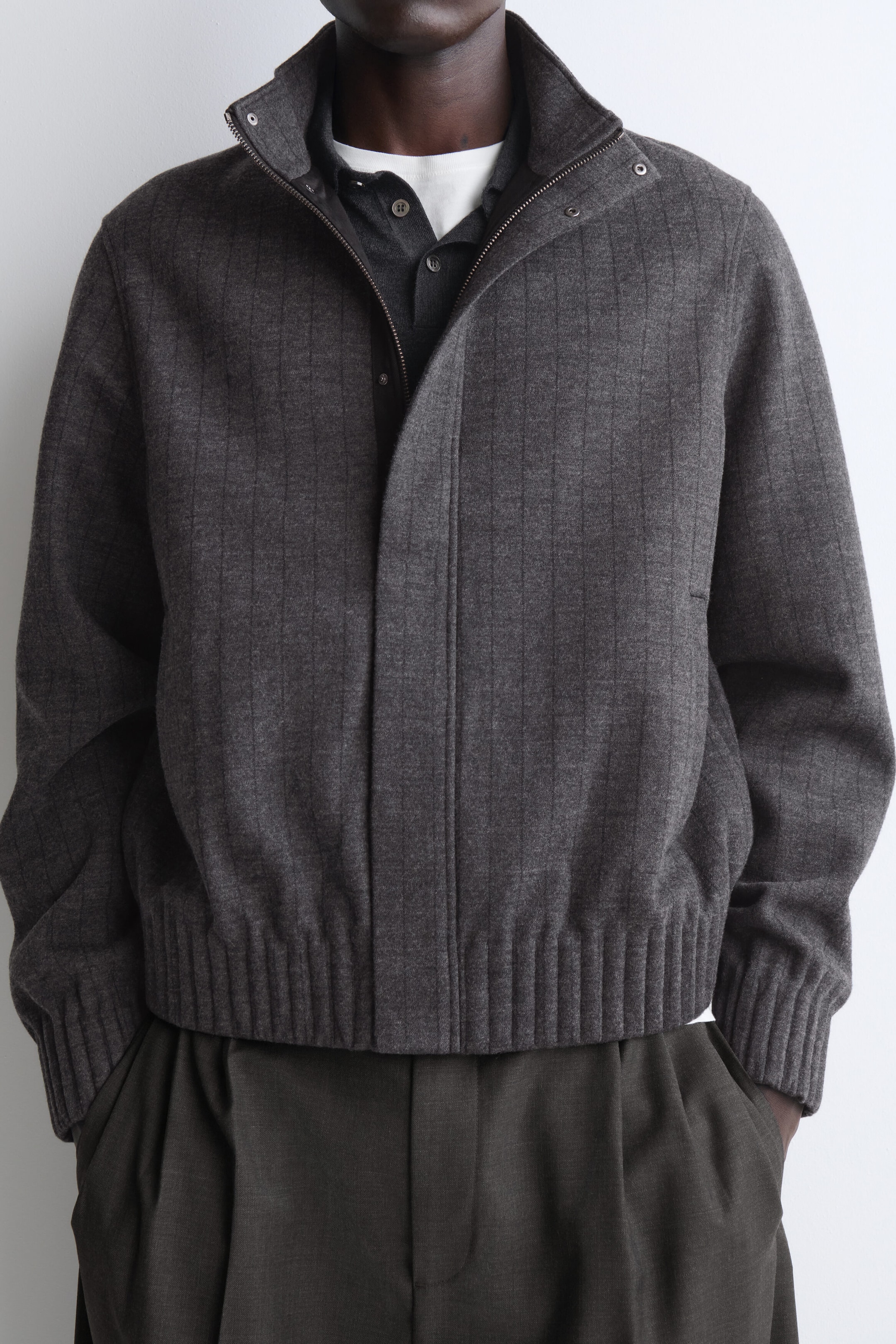 A top-half view shows a dark gray bomber jacket featuring subtle vertical ribbing, a high stand collar, and ribbed hem and cuffs. The jacket is worn layered over a dark gray button-down shirt and a white T-shirt, highlighting its structured, relaxed fit. A top-half view shows a dark gray bomber jacket featuring subtle vertical ribbing, a high stand collar, and ribbed hem and cuffs. The jacket is worn layered over a dark gray button-down shirt and a white T-shirt, highlighting its structured, relaxed fit.