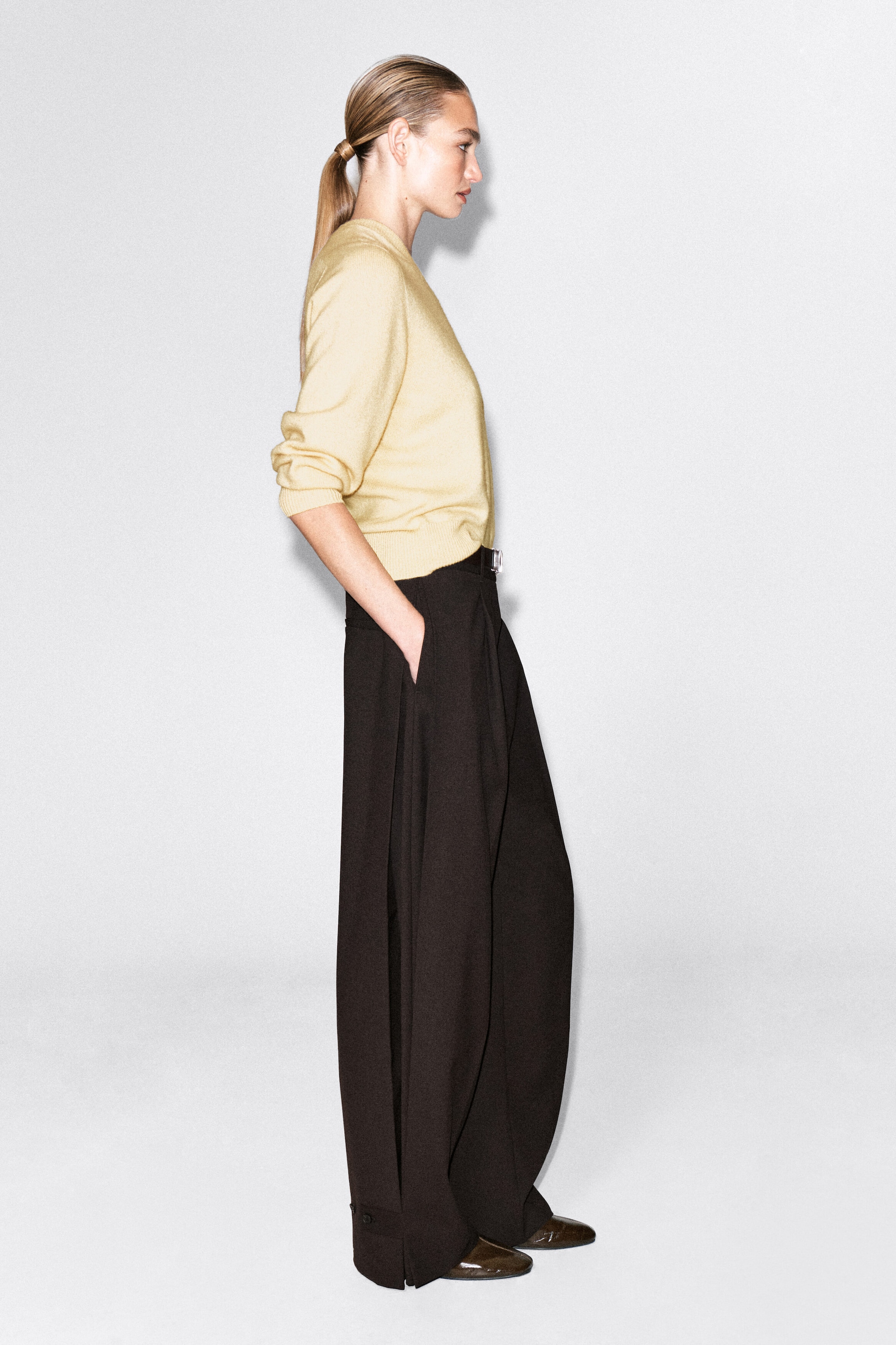 Ein hellgelber Strickpullover mit Rundhalsausschnitt und dunkelbraune Wide-Leg-Hose werden von dem Modell in Profilansicht getragen. Der Pullover verfügt über gerippte Bündchen und einen gerippten Saum, während die hoch taillierte Hose mit einer Hand in der Seitentasche gestylt ist und Seitenschlitze mit Knopfdetails am Saum aufweist. Ein hellgelber Strickpullover mit Rundhalsausschnitt und dunkelbraune Wide-Leg-Hose werden von dem Modell in Profilansicht getragen. Der Pullover verfügt über gerippte Bündchen und einen gerippten Saum, während die hoch taillierte Hose mit einer Hand in der Seitentasche gestylt ist und Seitenschlitze mit Knopfdetails am Saum aufweist.
